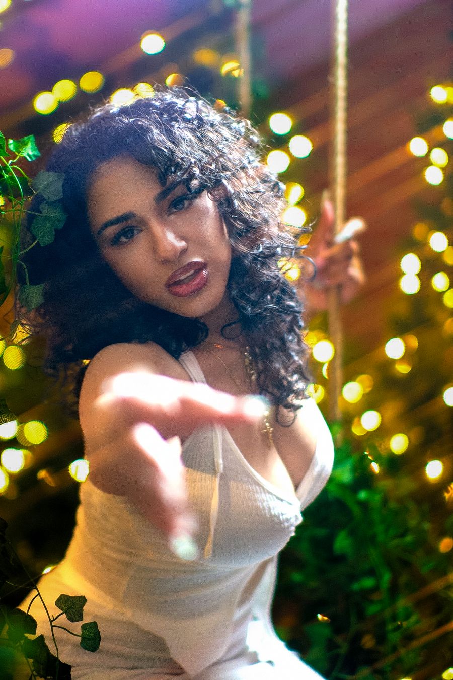 Young woman with curly hair in white dress reaching toward camera, surrounded by bokeh lights and green ivy.