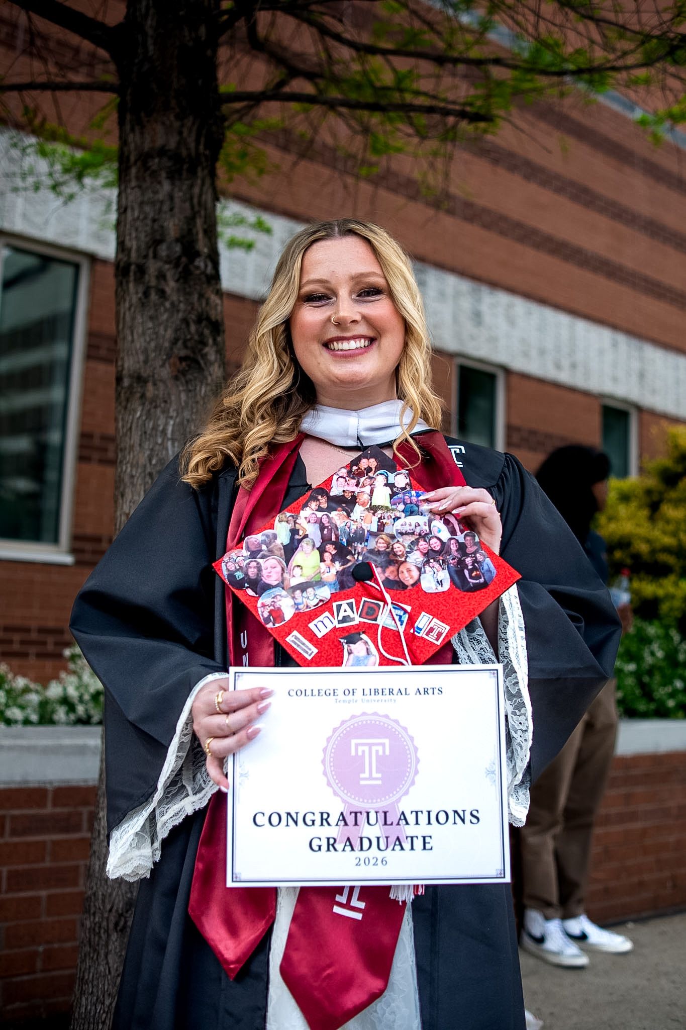 Temple University College of Liberal Arts graduate smiling in cap and gown holding congratulations sign outdoors.