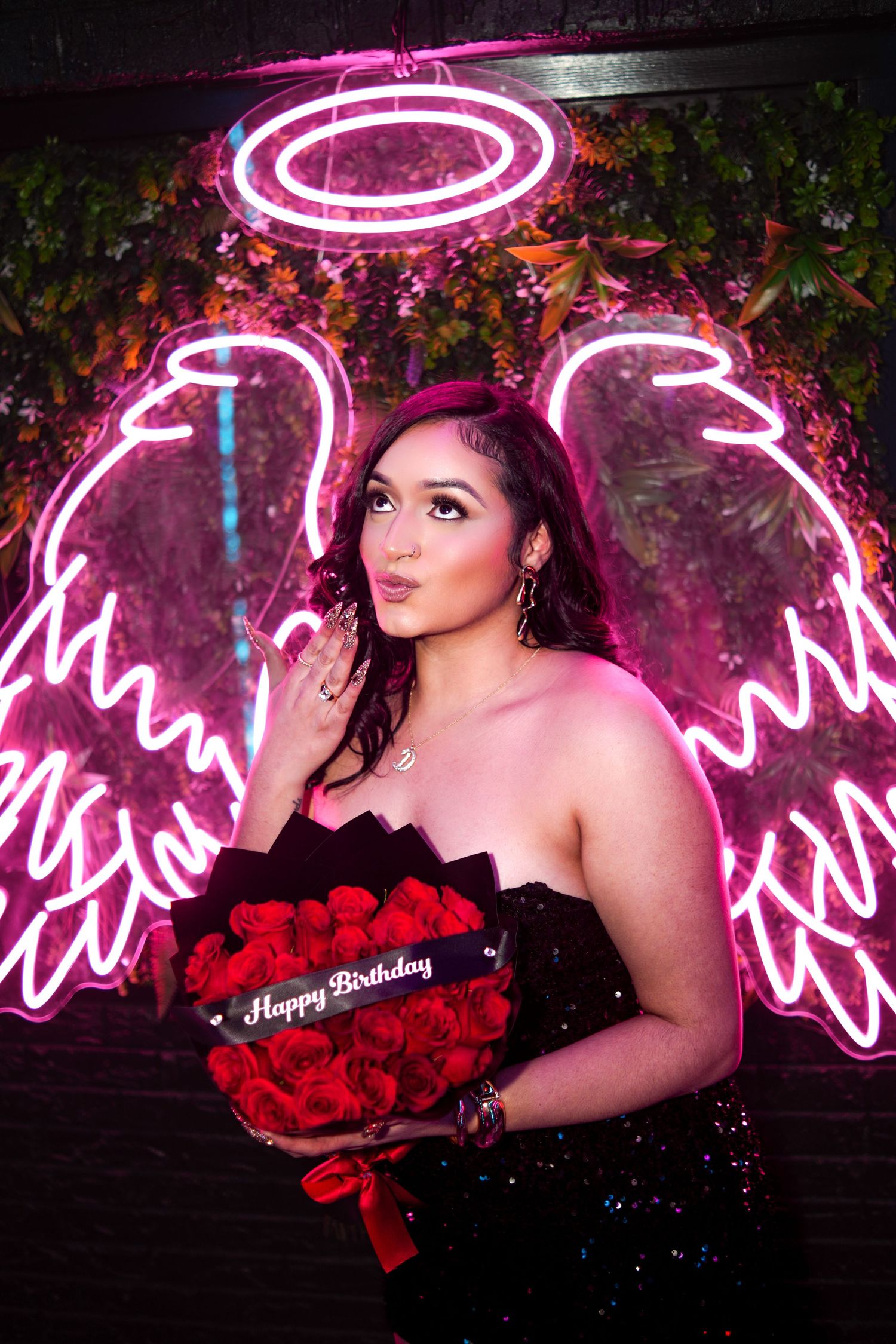 Woman in black strapless dress holds Happy Birthday red roses bouquet in front of pink neon angel wings.