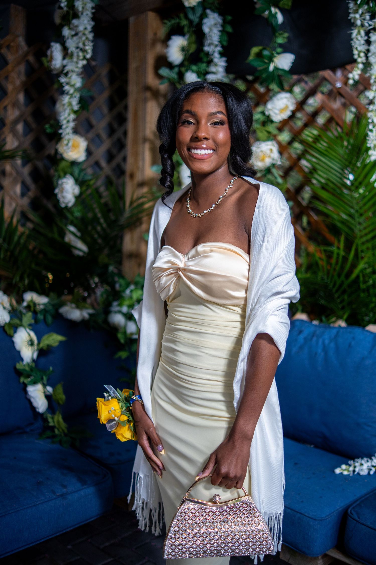 Smiling young woman in elegant cream satin dress with white shawl, holding bouquet and clutch purse at floral backdrop.