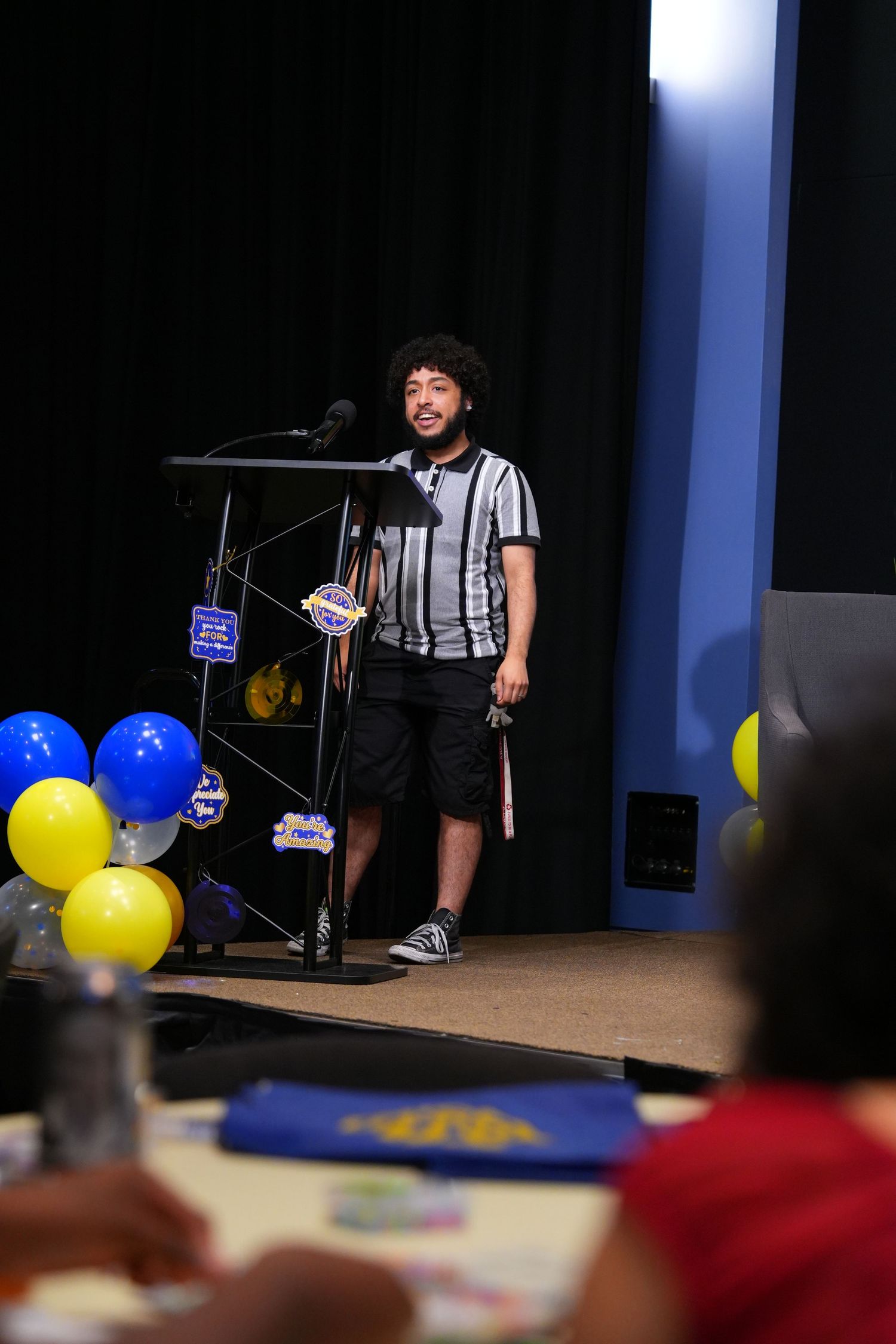Young man speaking at a podium with blue and yellow balloons at an indoor event or ceremony.
