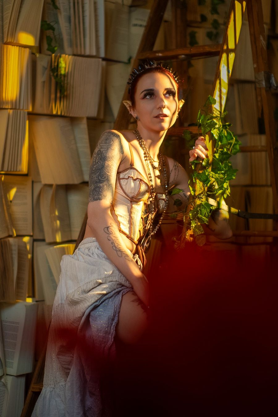 A tattooed woman in an elf costume with pointed ears holds greenery in a warmly lit fantasy book-lined setting.