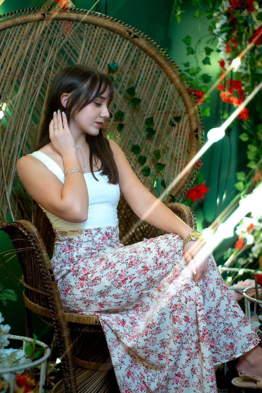 Young woman in floral maxi skirt sitting in wicker peacock chair surrounded by lush green plants and flowers.