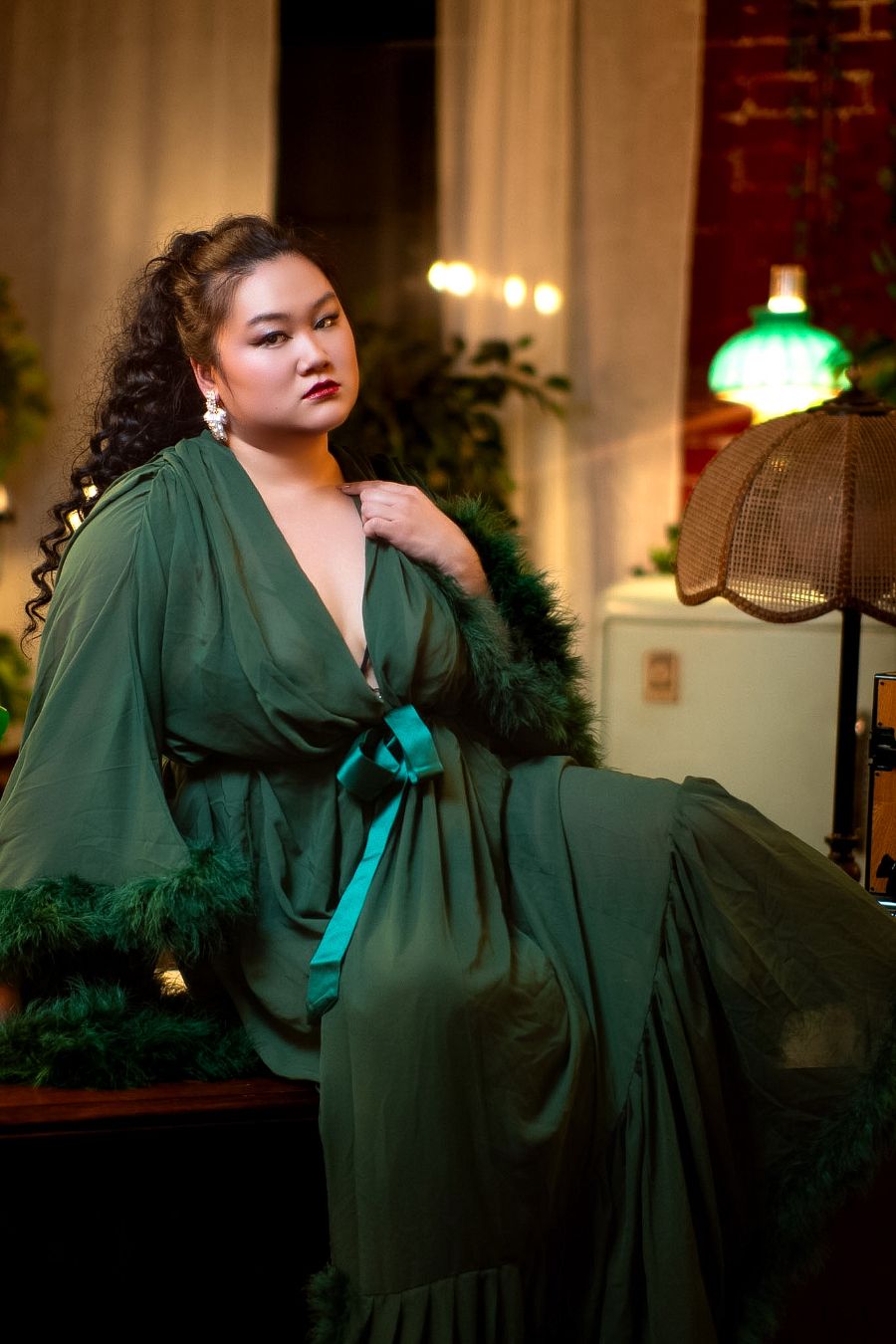 Woman in elegant green satin wrap gown with feather trim sits gracefully in a warmly lit, plant-filled room.