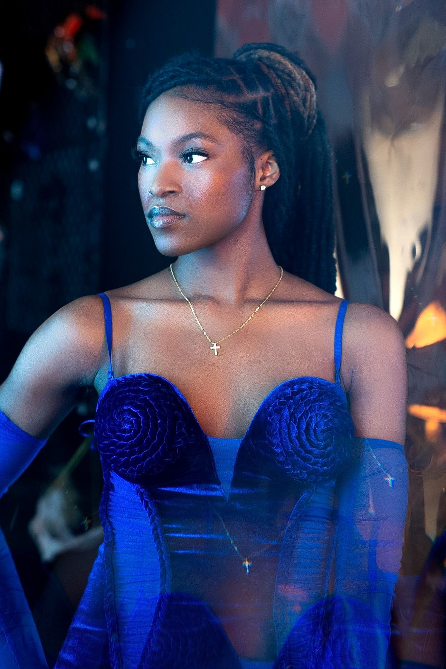 Young Black woman in blue velvet corset gown with cross necklace poses elegantly under blue lighting.