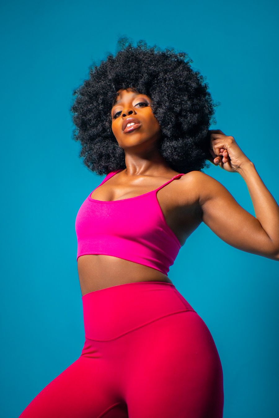 Black woman with afro in pink crop top and leggings posing confidently against a blue background.