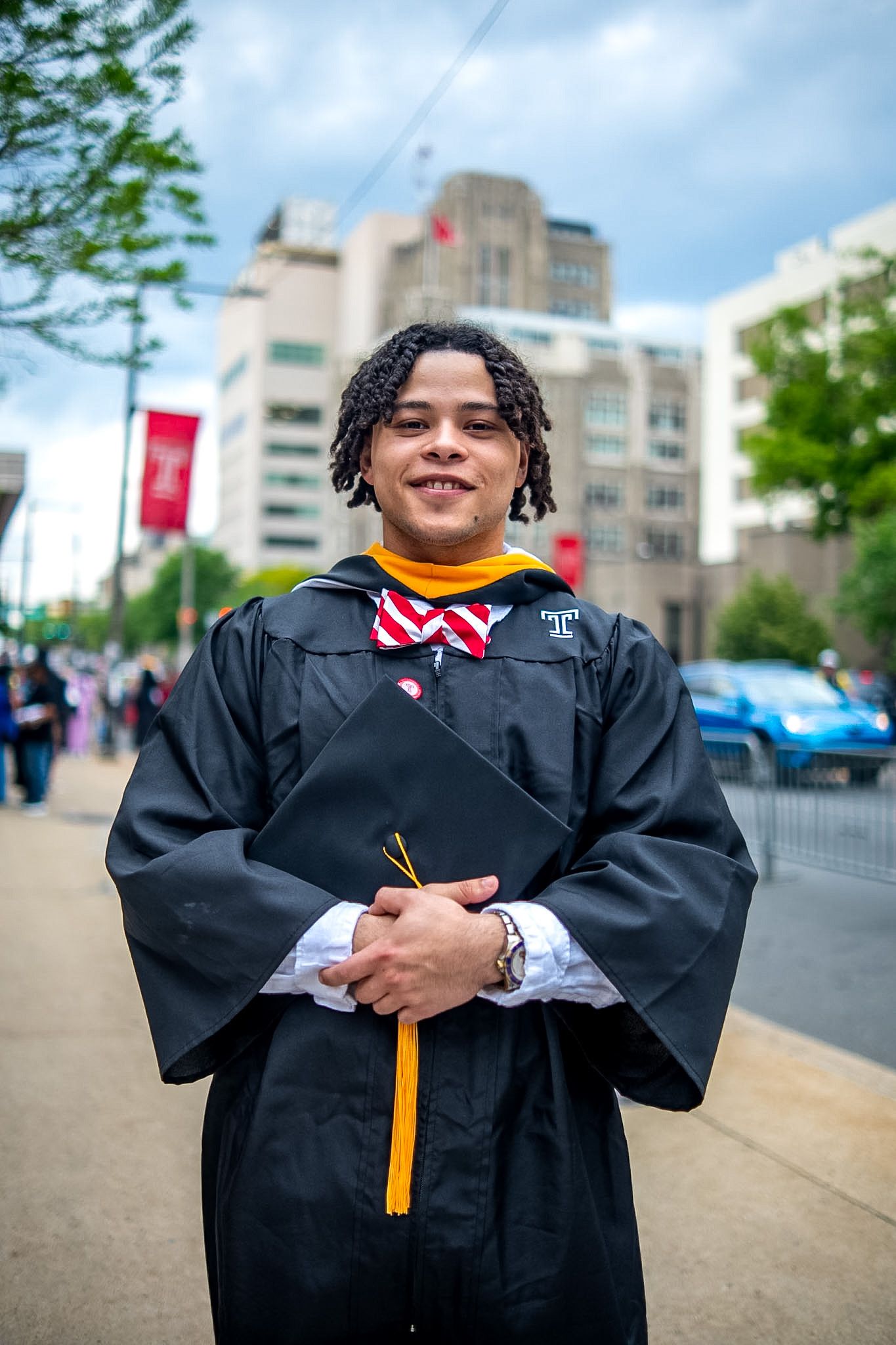 Young man in black graduation gown with gold hood and red bow tie smiling outdoors at Temple University commencement.