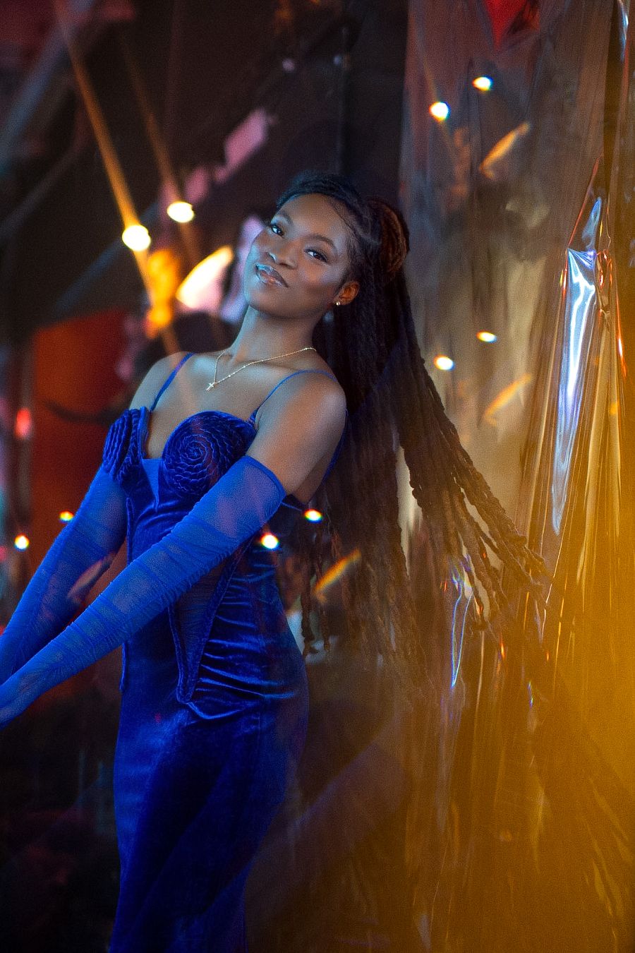 A young woman in a blue velvet gown and long gloves poses elegantly under colorful lights at a glamorous event.