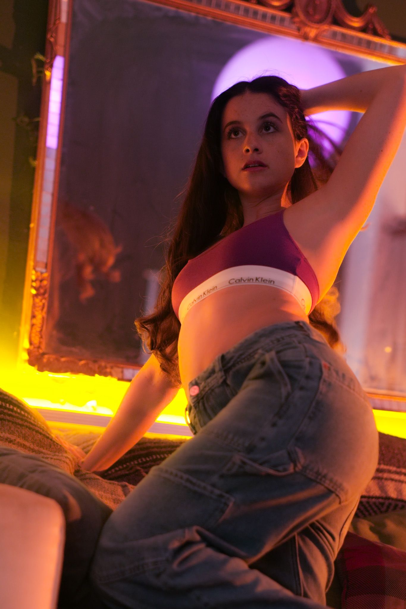 Young woman in Calvin Klein bralette and jeans posing by ornate mirror with purple neon lighting.