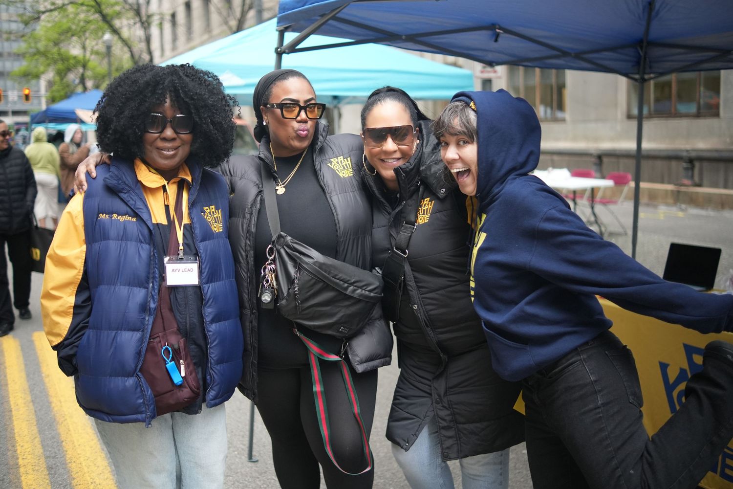 Four young women in school jackets smile and pose together at an outdoor community event.