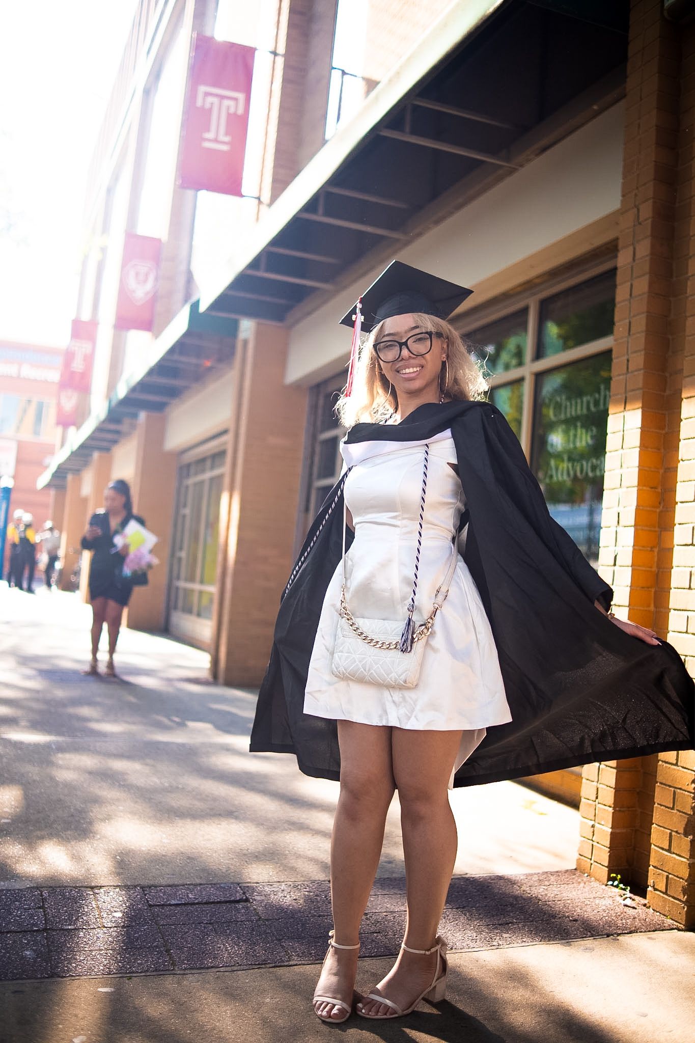 Young woman in graduation cap and gown smiles on a sunny campus sidewalk wearing a white dress and heels.