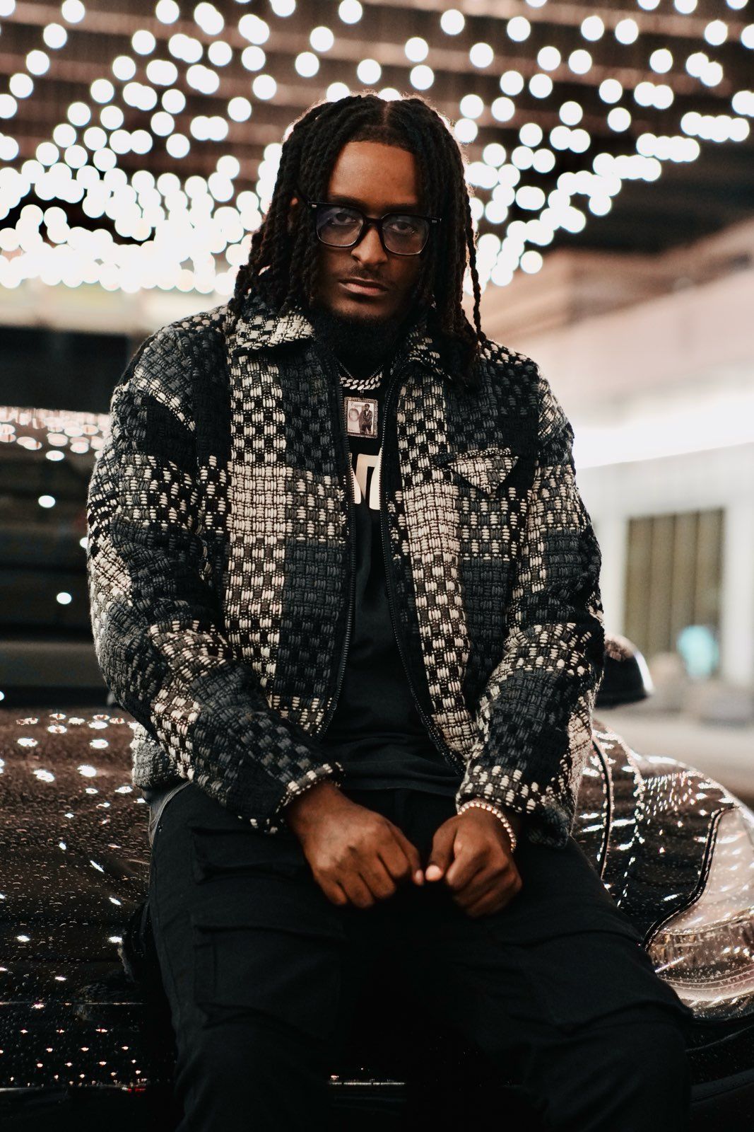 Stylish man with braids wearing patterned jacket and glasses sits under bokeh lights at night.