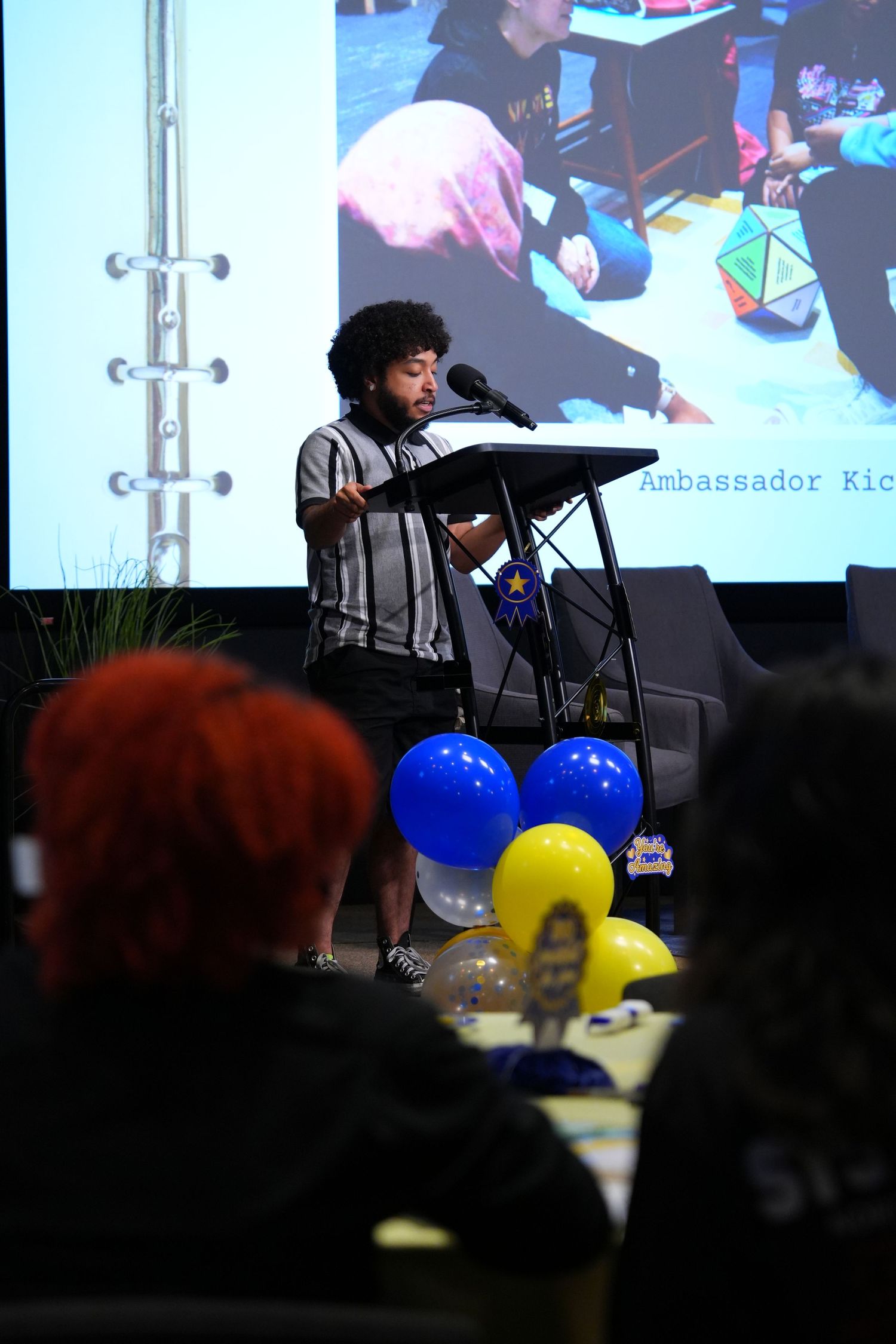 A speaker presents at a podium during an Ambassador Kickoff event with blue and yellow balloons in the foreground.