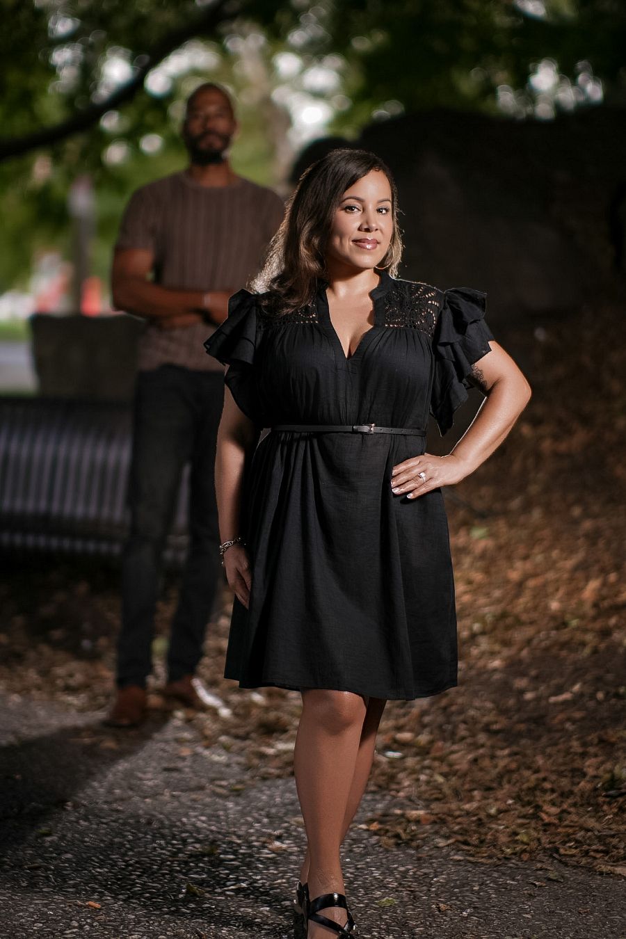 Woman in black dress poses confidently outdoors with man in brown shirt standing in background.