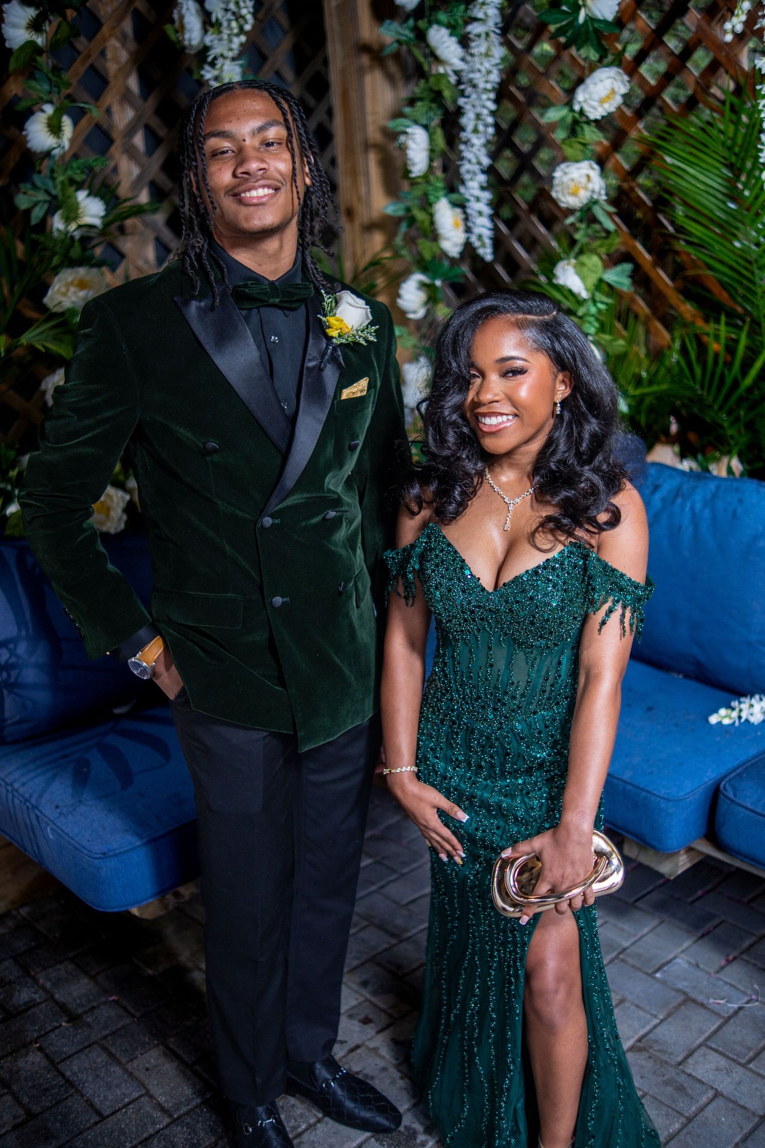 A couple dressed in matching emerald green formal attire poses together at prom in front of a floral backdrop.