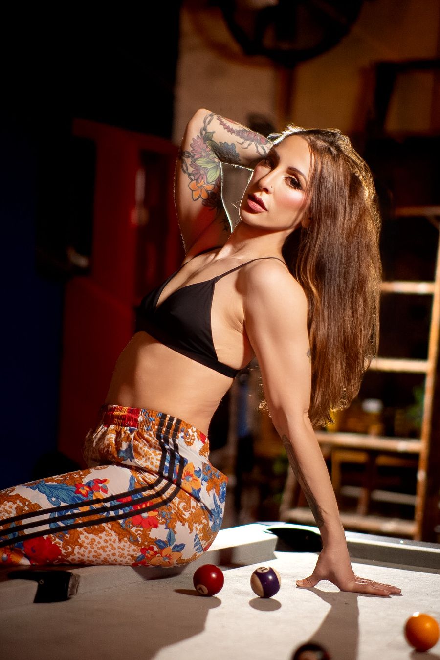Tattooed woman in black crop top and floral pants posing on a pool table in a dimly lit room.