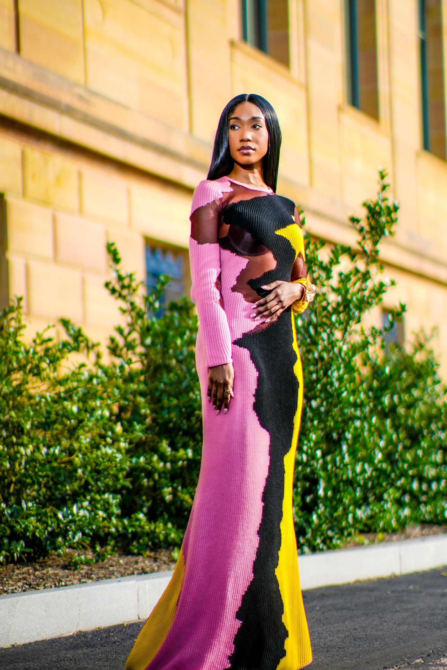 A Black woman poses outdoors in a stunning colorful pink, black, and yellow long-sleeve maxi dress near green shrubs.