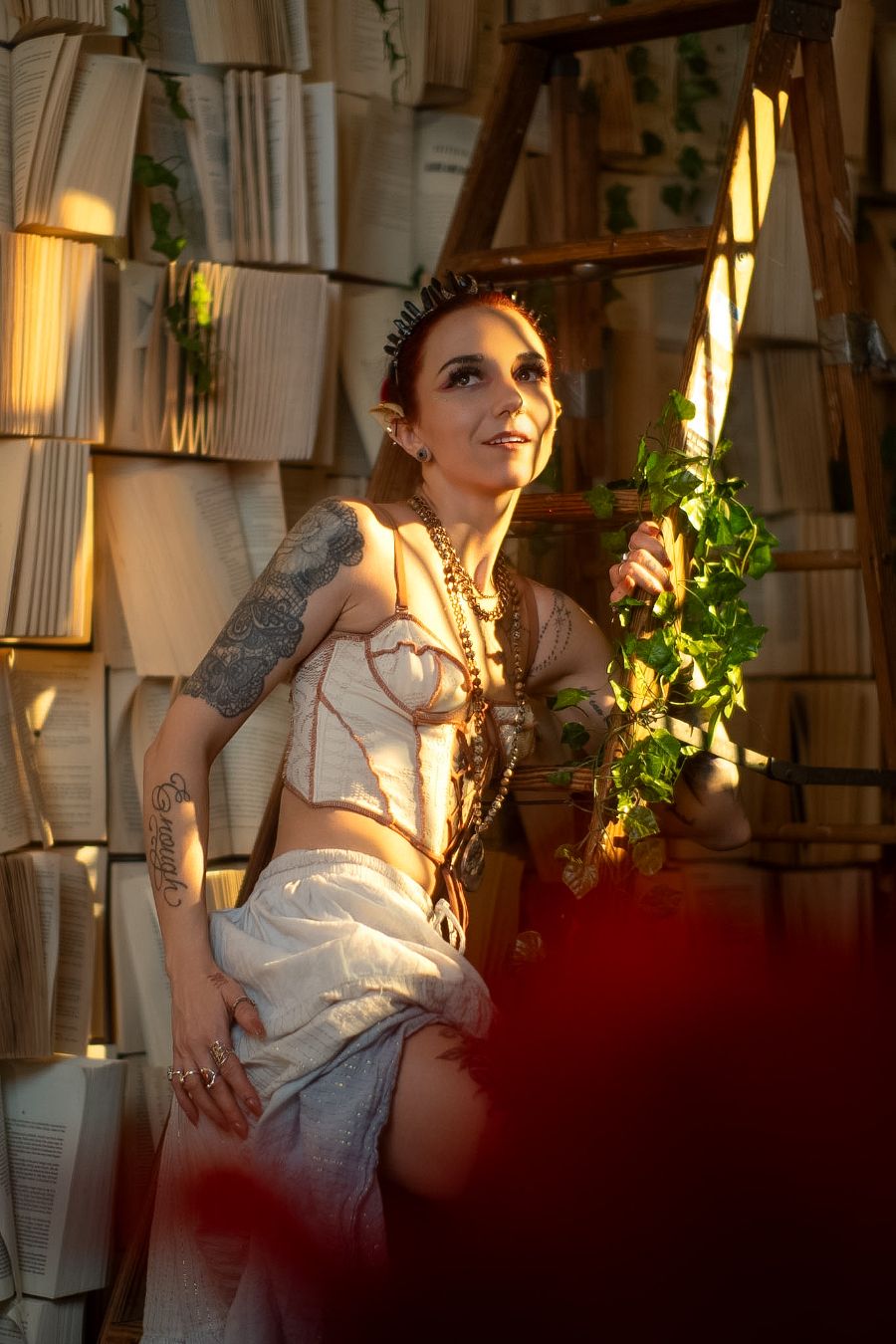 Tattooed woman in white corset top holding green vines, posed against wall of stacked books in warm golden light.