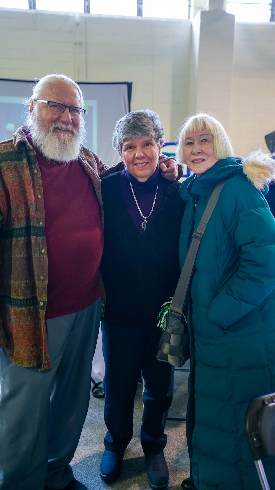 Three smiling adults posing together at an indoor event in winter clothing.