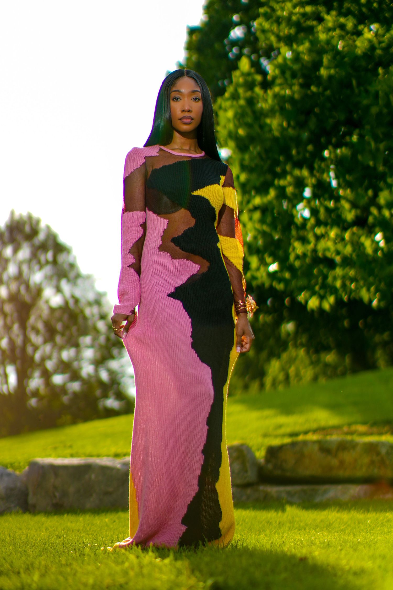 Tall woman in colorful abstract pink yellow black maxi dress stands on green grass near trees.