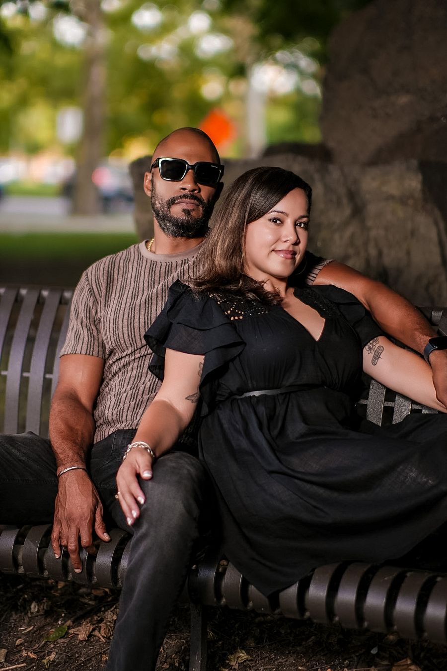 A couple sits together on a park bench, man in sunglasses and striped shirt, woman in black dress, outdoors.