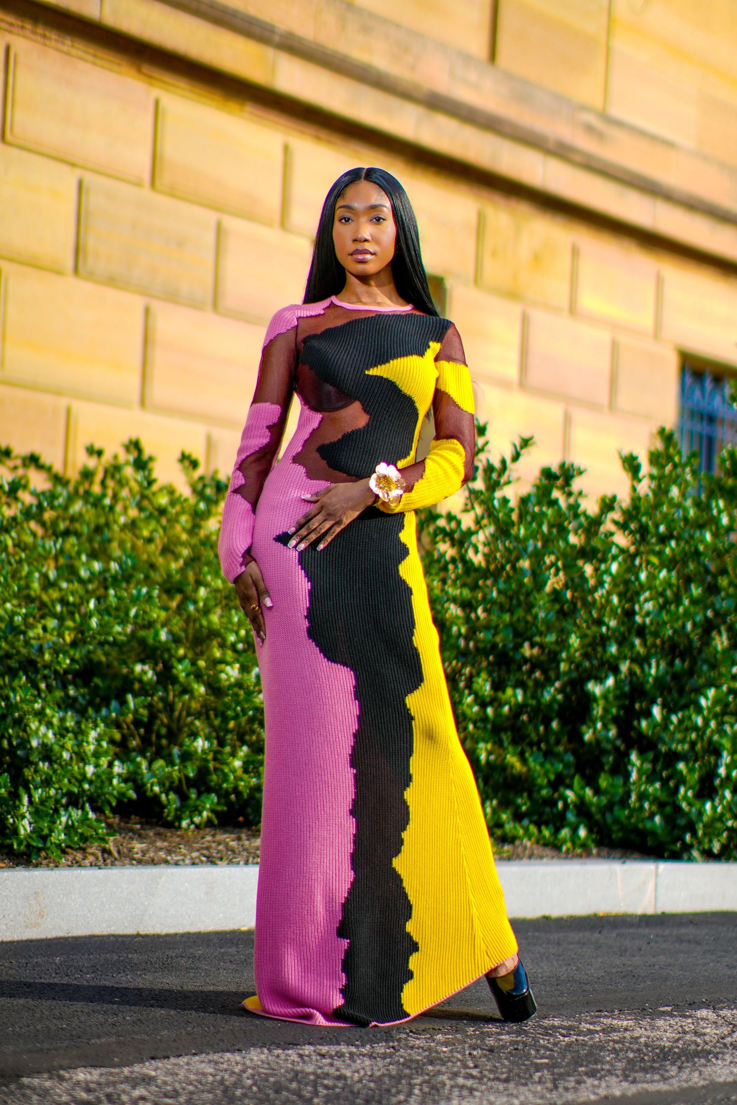 Woman in vibrant pink yellow black abstract ribbed maxi dress poses against golden stone wall outdoors.