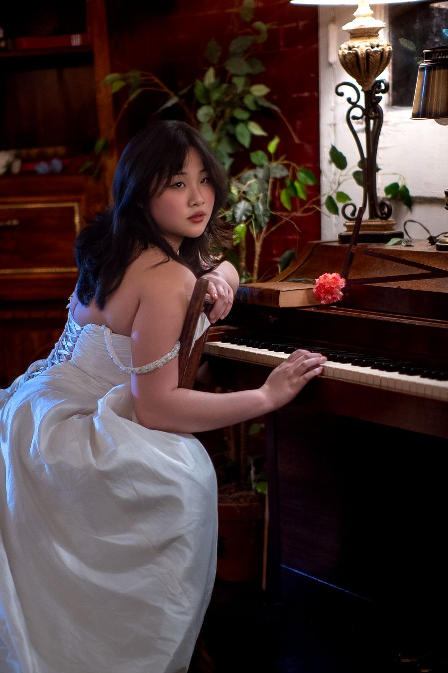 Young woman in white gown playing an upright piano in a warmly lit vintage room with plants and decorative lamp.