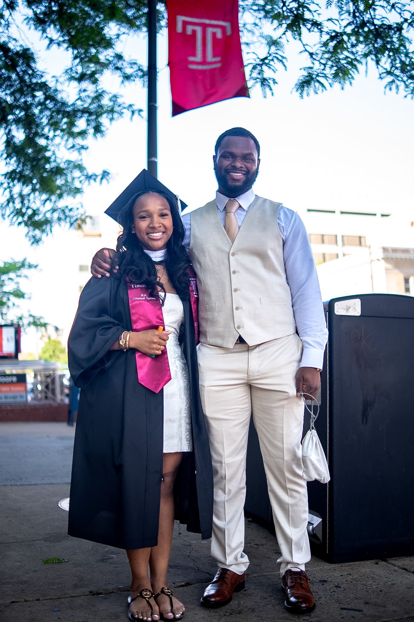 Temple University graduate in cap and gown poses with a man in a beige suit near a Temple T banner.