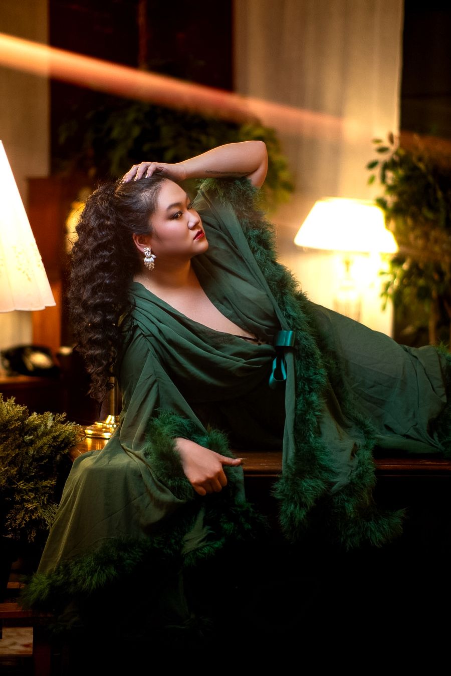 A woman in an elegant green feathered gown poses gracefully in a warmly lit, luxurious room.