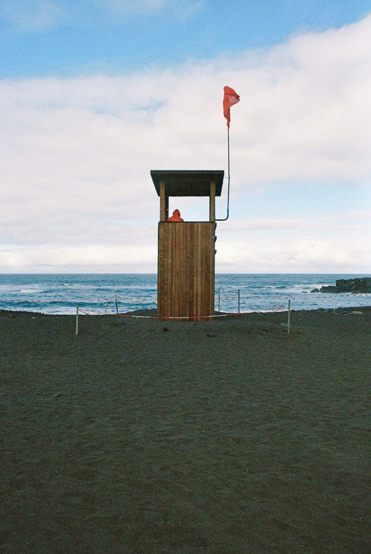 Wooden lifeguard tower with red flag on black sand beach overlooking the ocean.