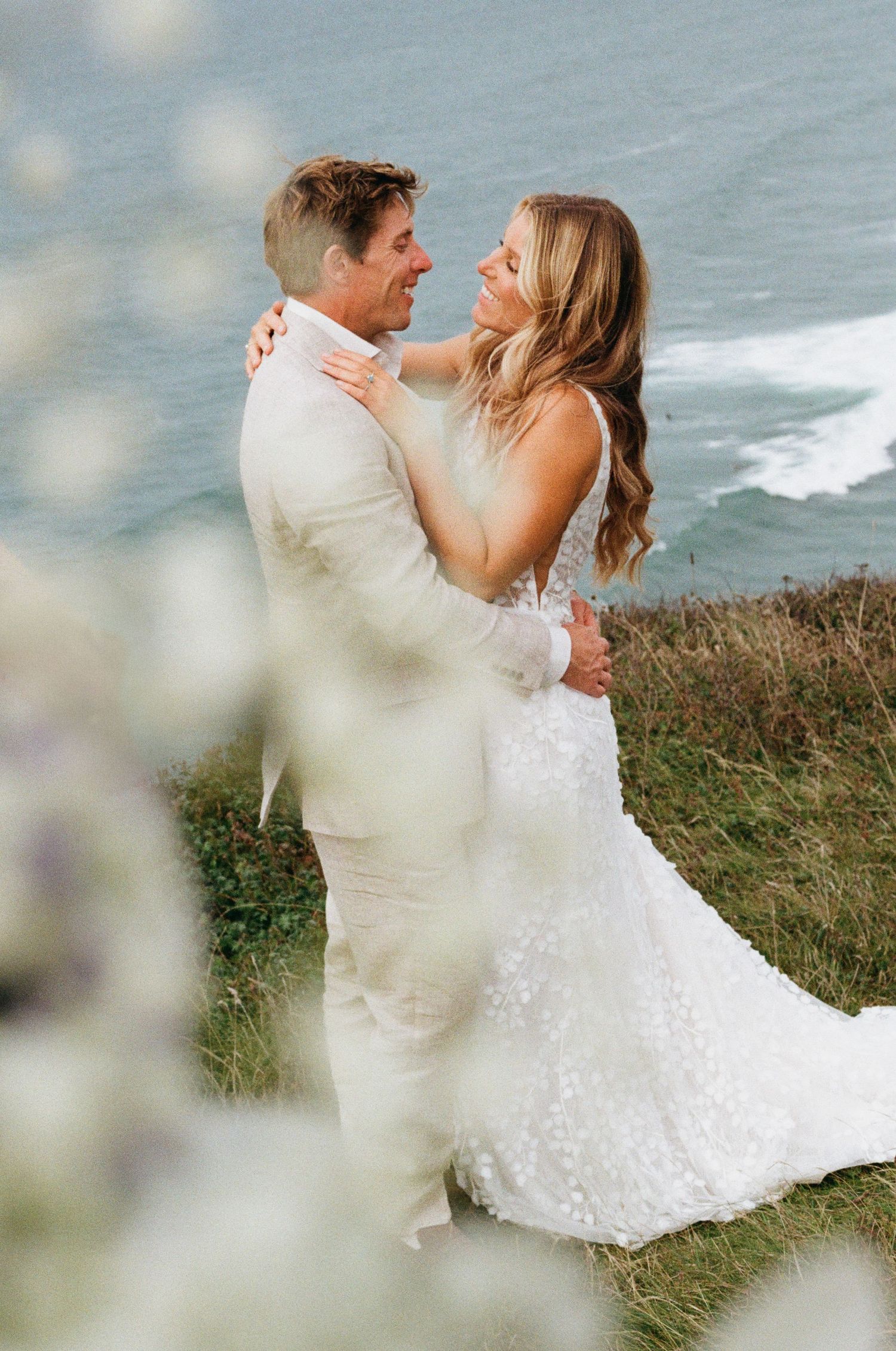 A romantic wedding couple embraces on a coastal cliff overlooking the ocean in dreamy lighting.