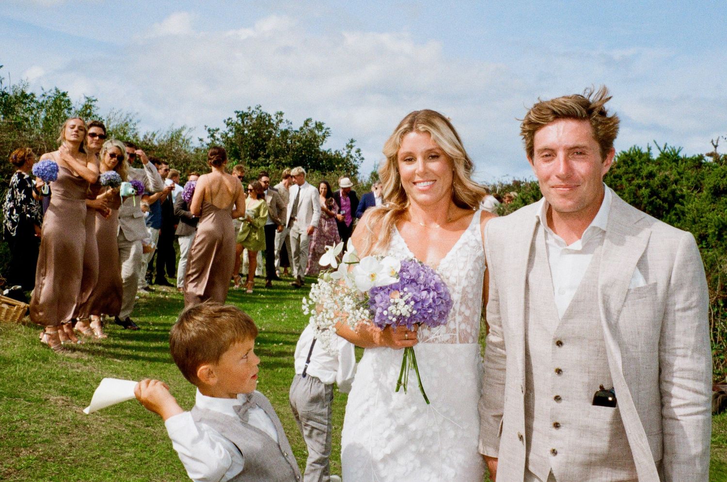 A wedding party celebrates on a sunny lawn with guests in summer attire.