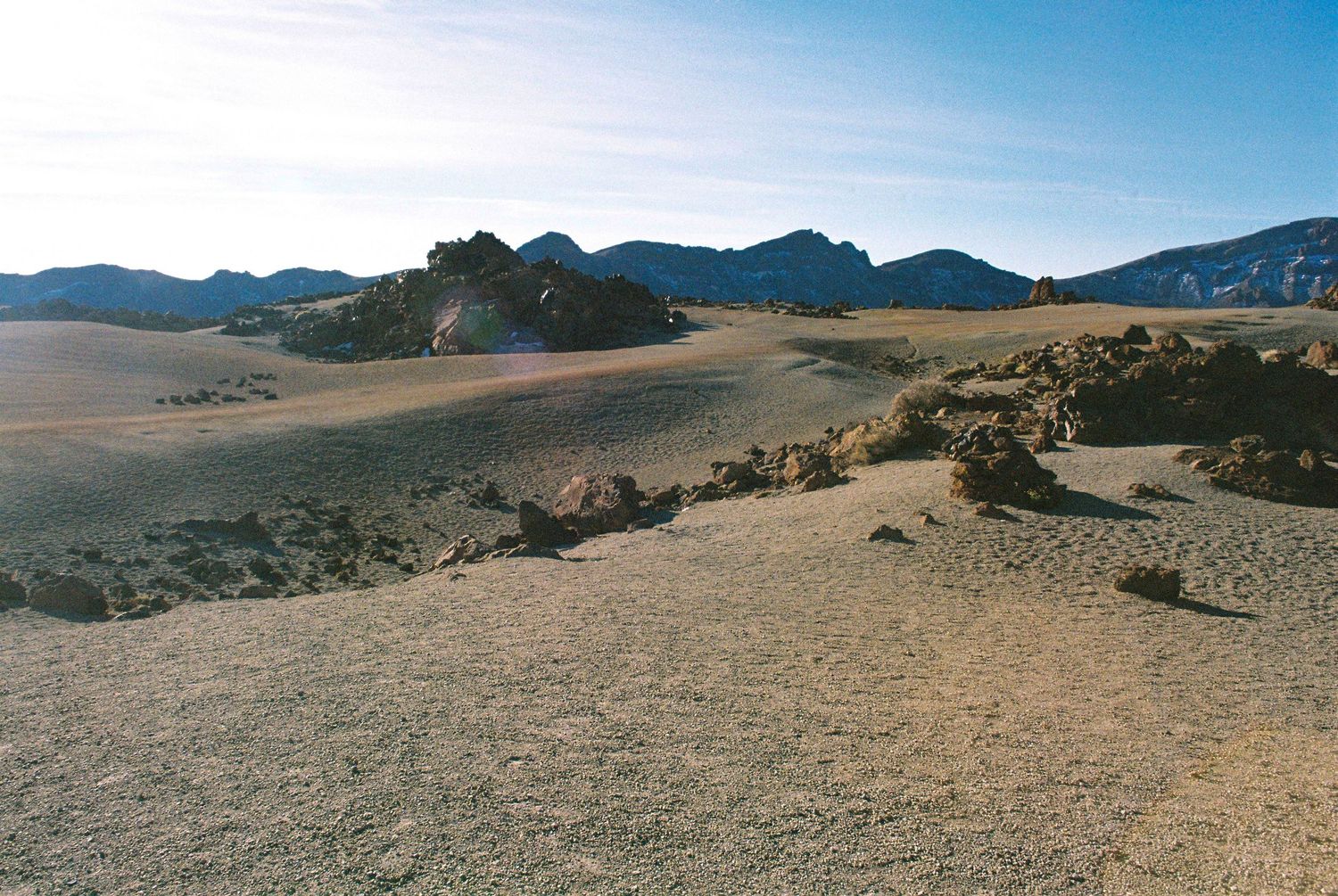 Panoramic view of a vast desert landscape with rolling sand dunes and distant mountains under a bright blue sky.