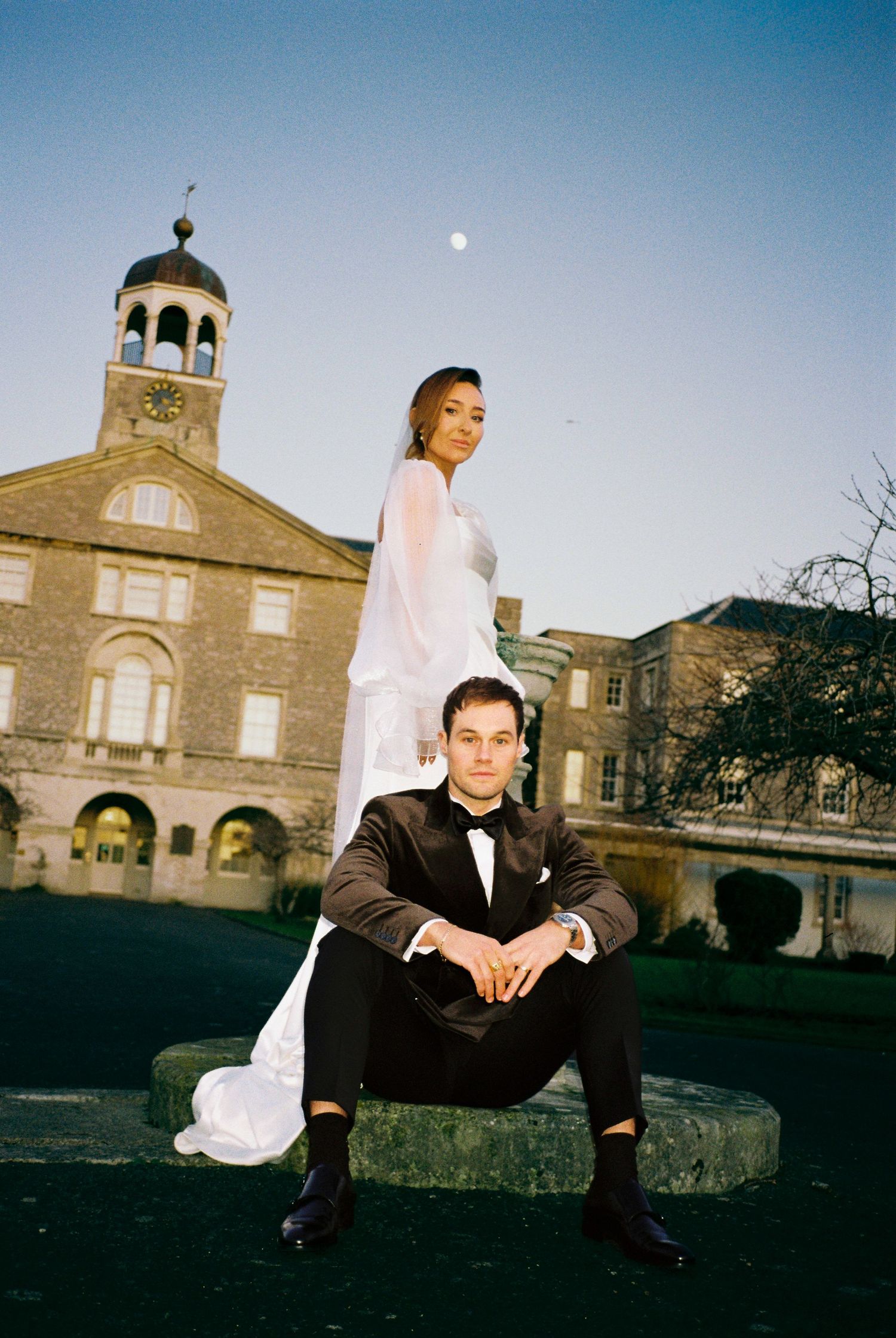 Person in formal attire sits on stone wall with historic building architecture in background.