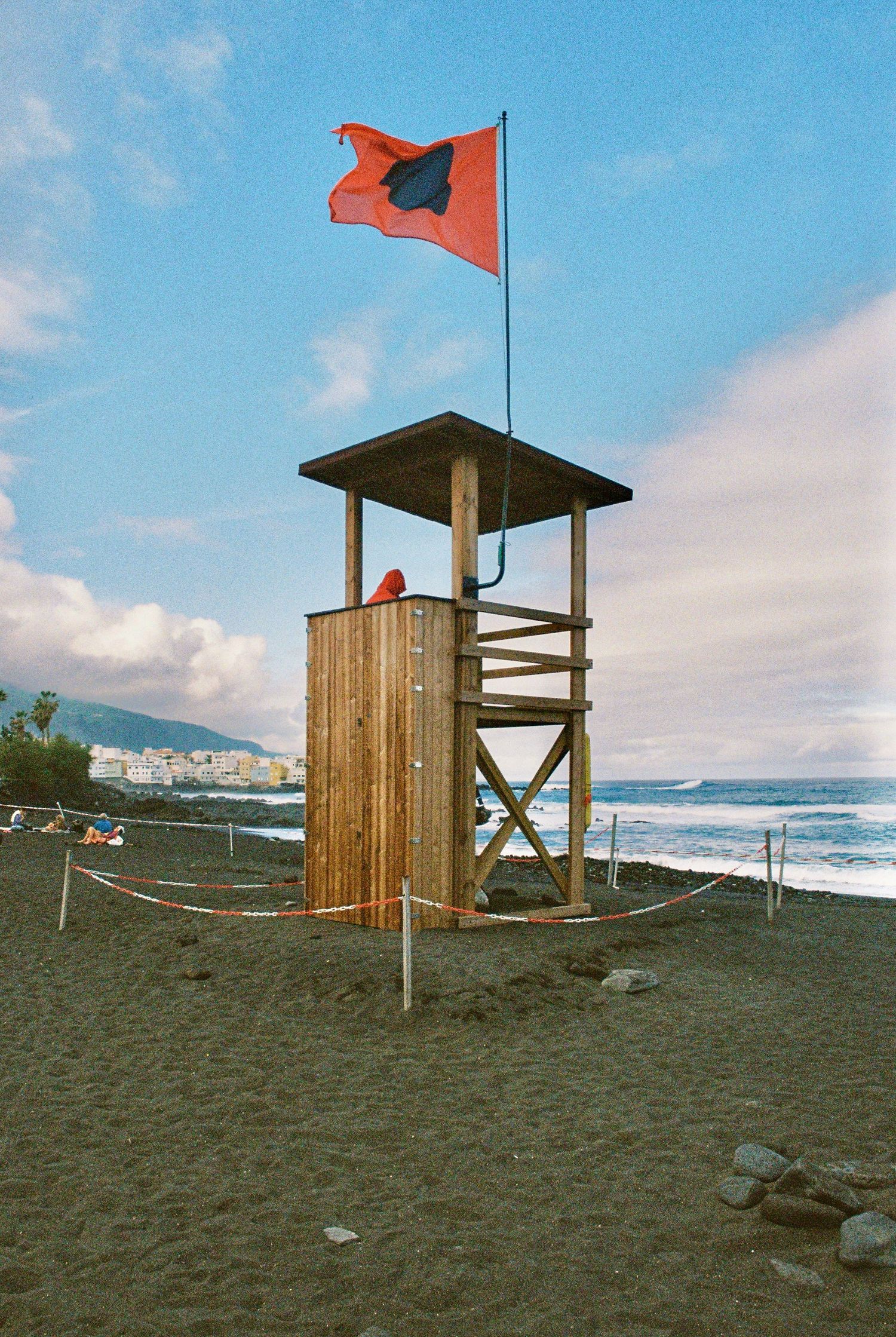 Wooden lifeguard tower with red flag on black sand beach overlooking ocean waves.