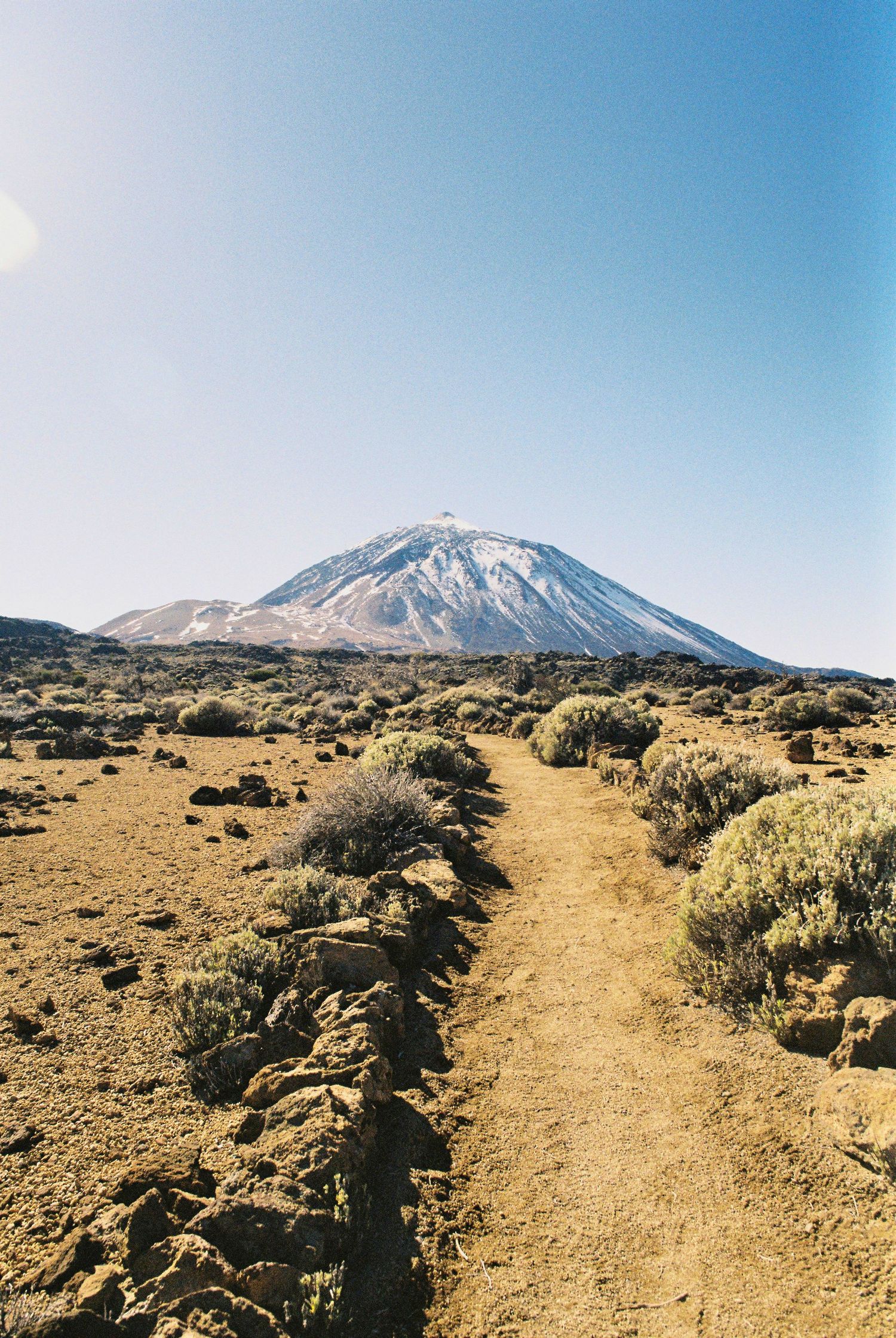 Desert hiking trail leading towards snow-capped Mount Teide through arid volcanic landscape.
