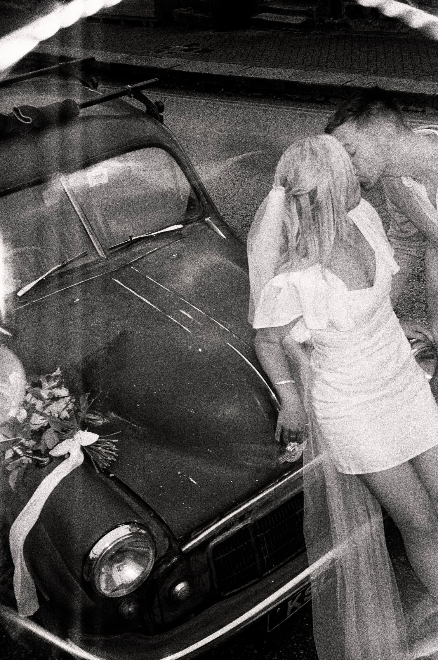 A couple in wedding attire poses near a classic black car.