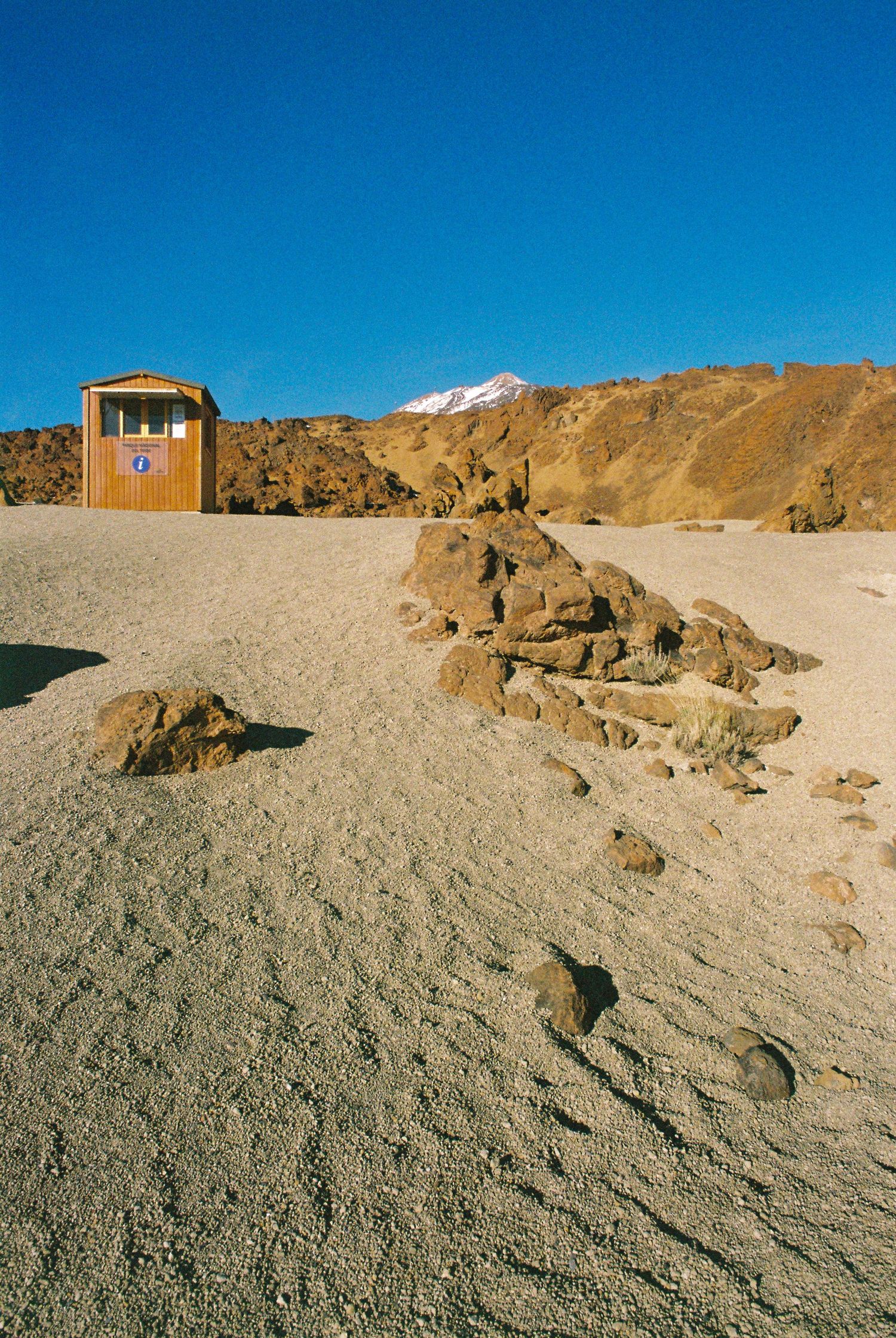 Rocky desert terrain with snow-capped mountains visible in the distance under a bright blue sky.