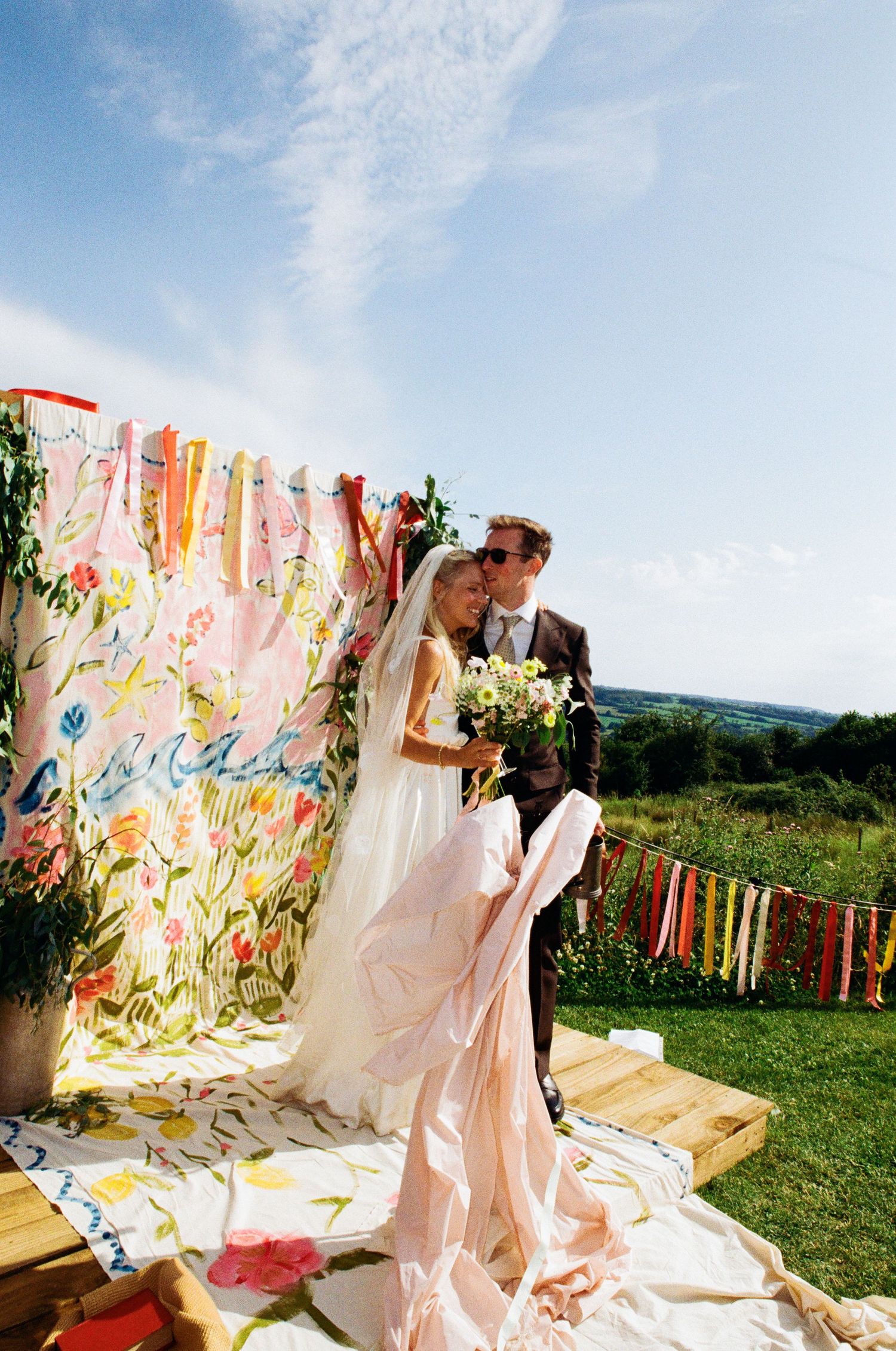 Bride and groom standing before a colorful floral backdrop with ribbon streamers at an outdoor wedding ceremony.