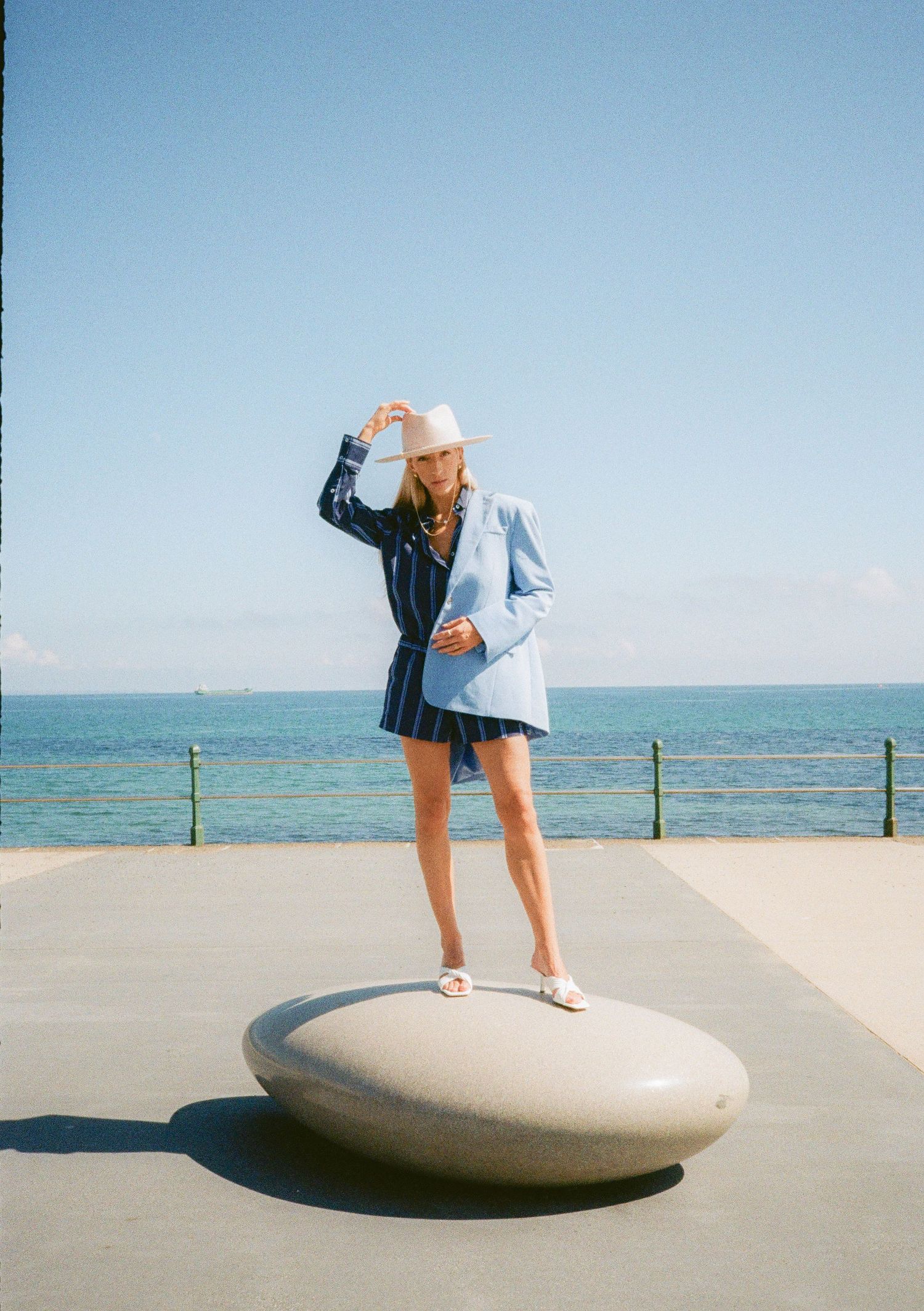 A person in casual attire poses playfully on a large stone sculpture near the beach.