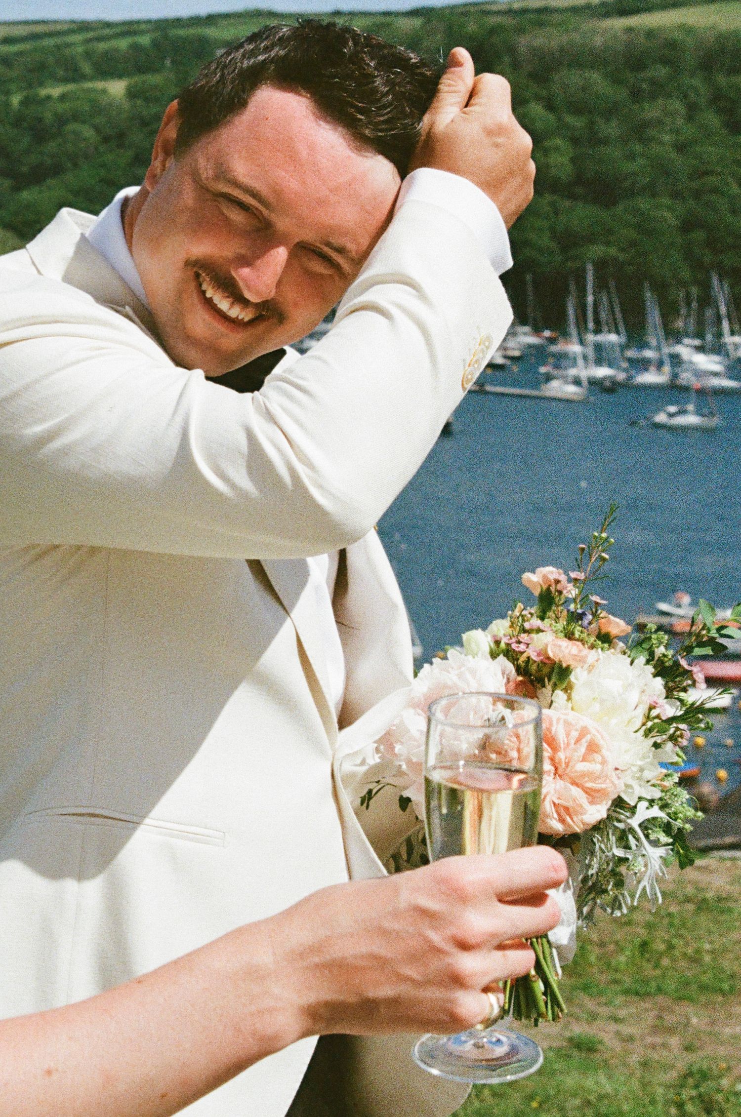 Wedding guest in white jacket toasts with champagne against marina backdrop.