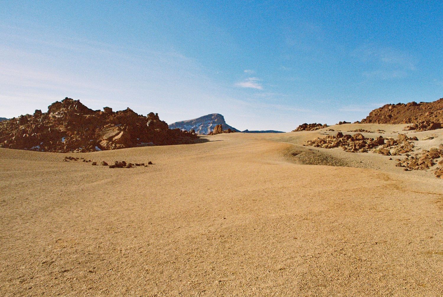 Rolling sand dunes and rocky outcrops stretch across an arid desert landscape under bright sunlight.