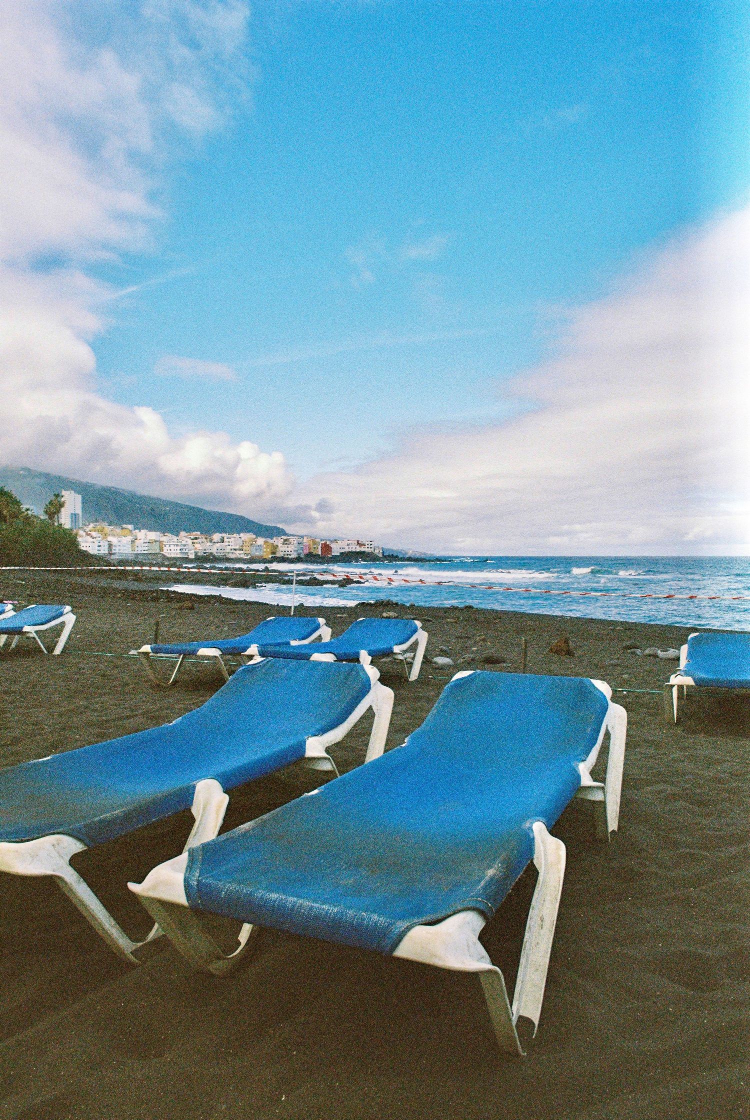 Empty blue lounge chairs on a black sand beach with mountains and ocean in the background.