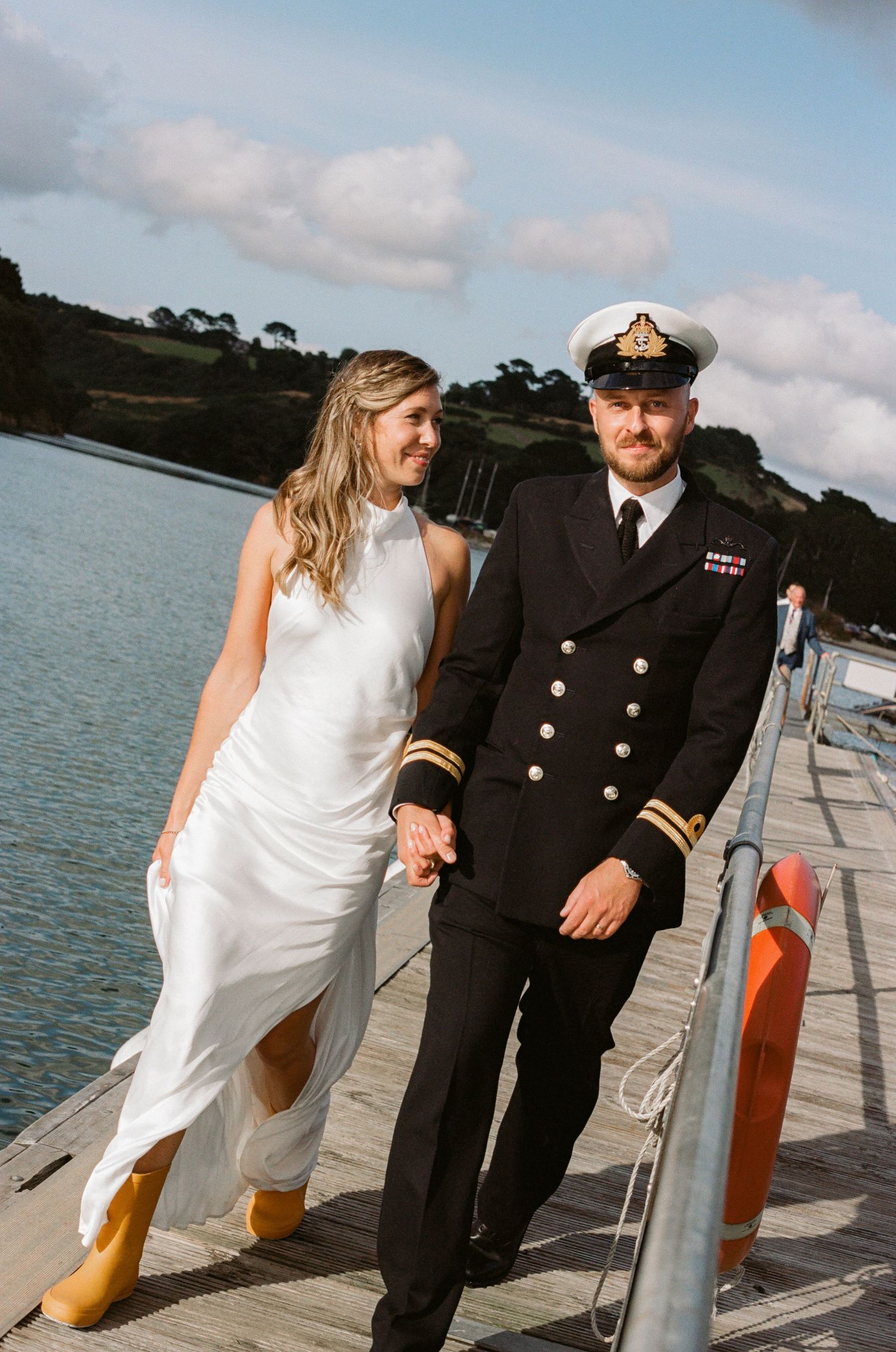 A couple walks along a waterfront dock, with the bride in white dress and groom in naval uniform.