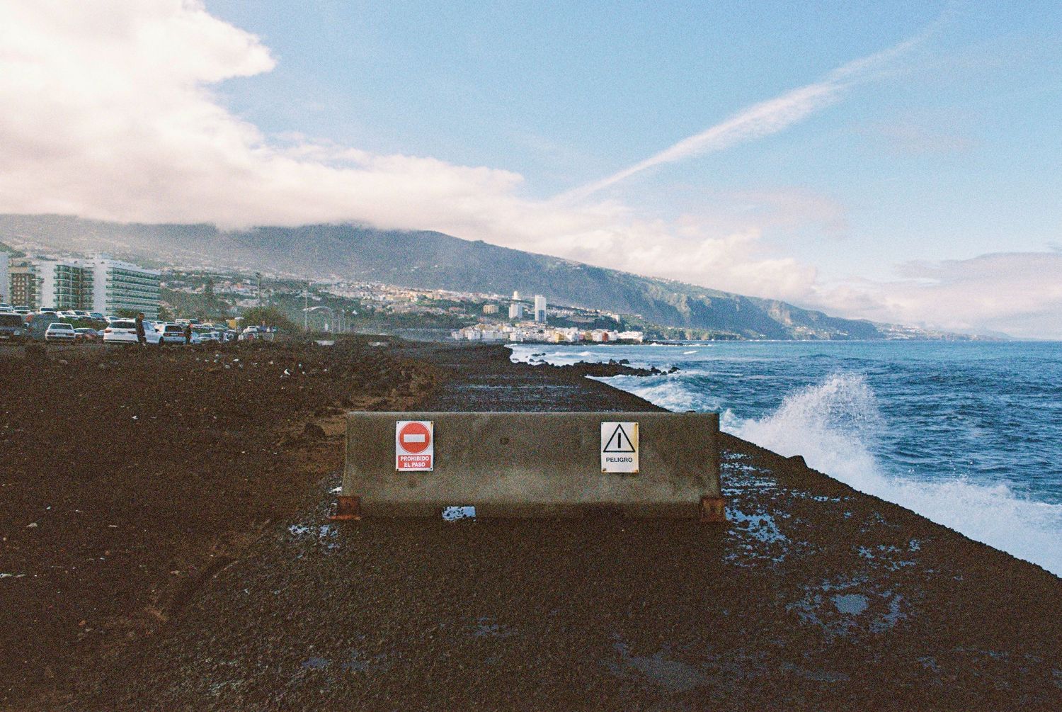 Warning signs stand on a rocky coast with crashing waves and mountains in the background.
