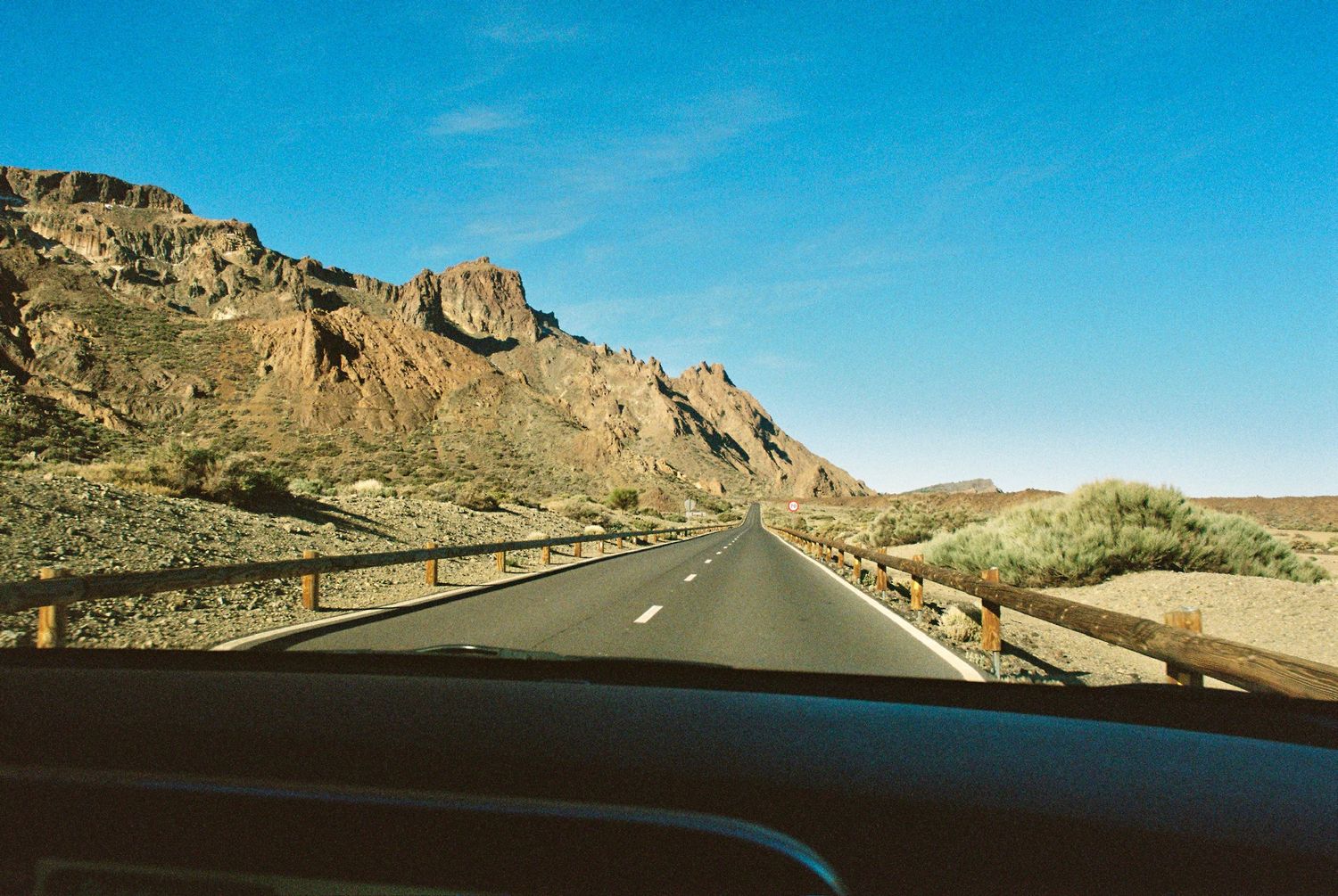 A desert highway stretches through rugged mountain terrain under clear blue skies.
