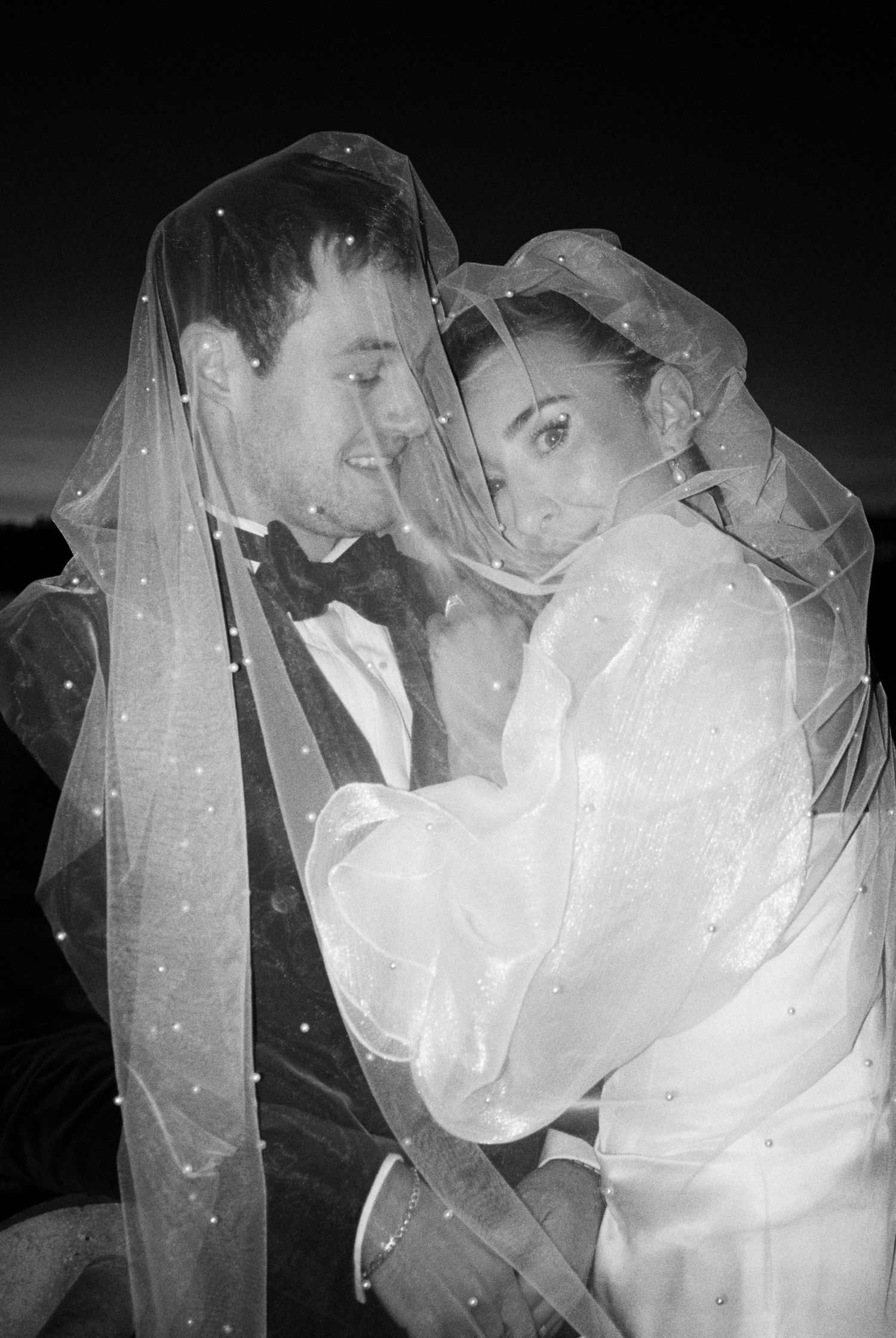 Black and white photo of romantic couple embracing under a flowing veil.