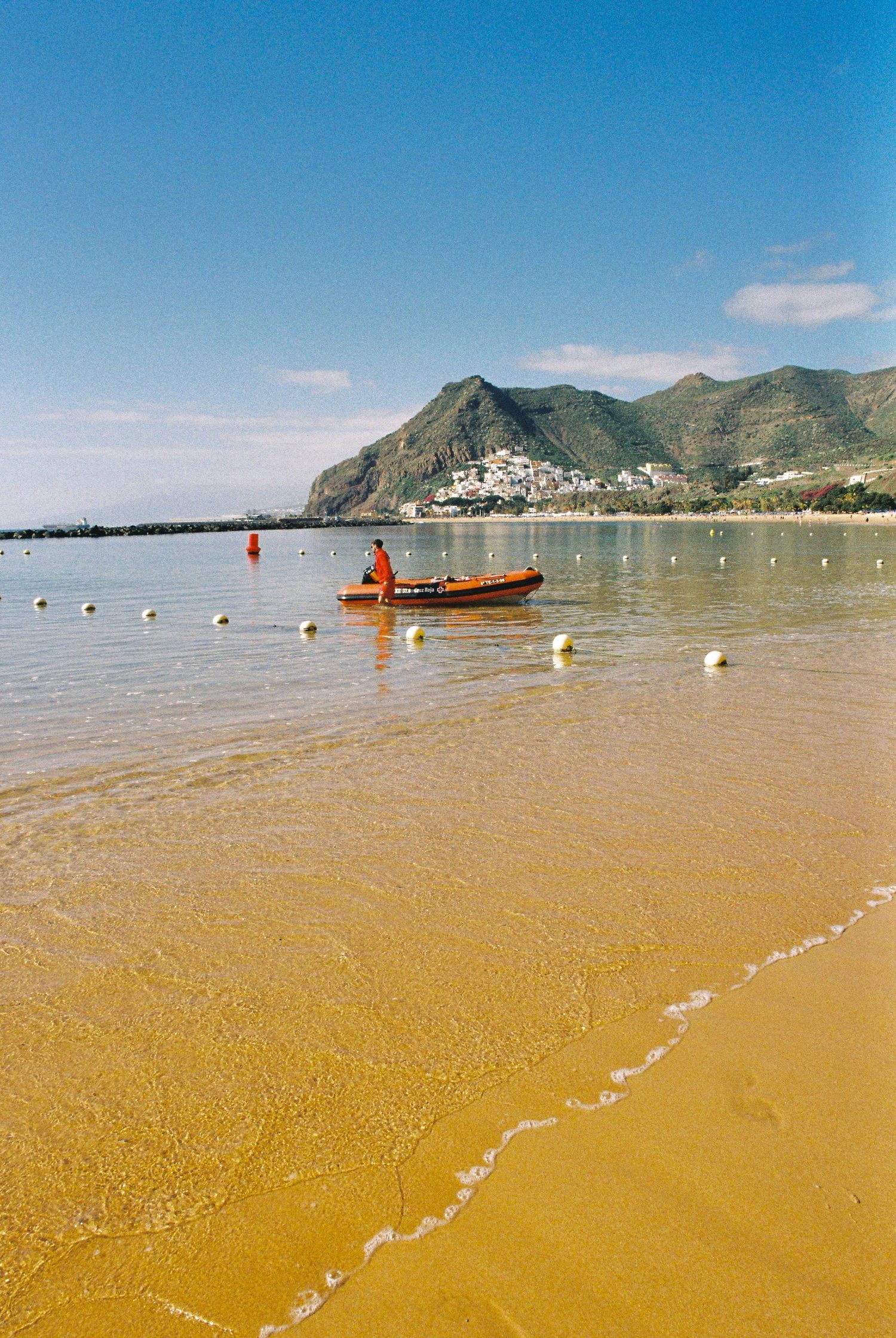 Sandy beach with yellow-tinted water stretches along mountainous coastline under blue sky.
