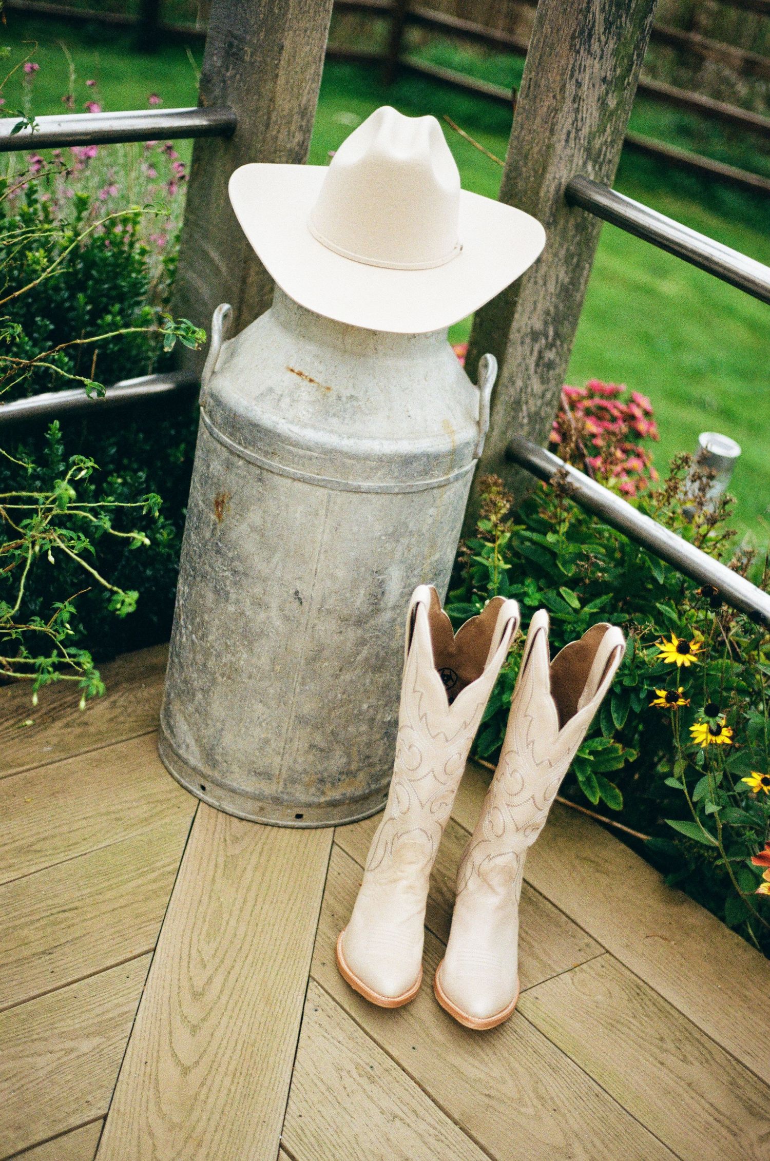 White cowboy boots and hat arranged near a rustic milk can on wooden deck.