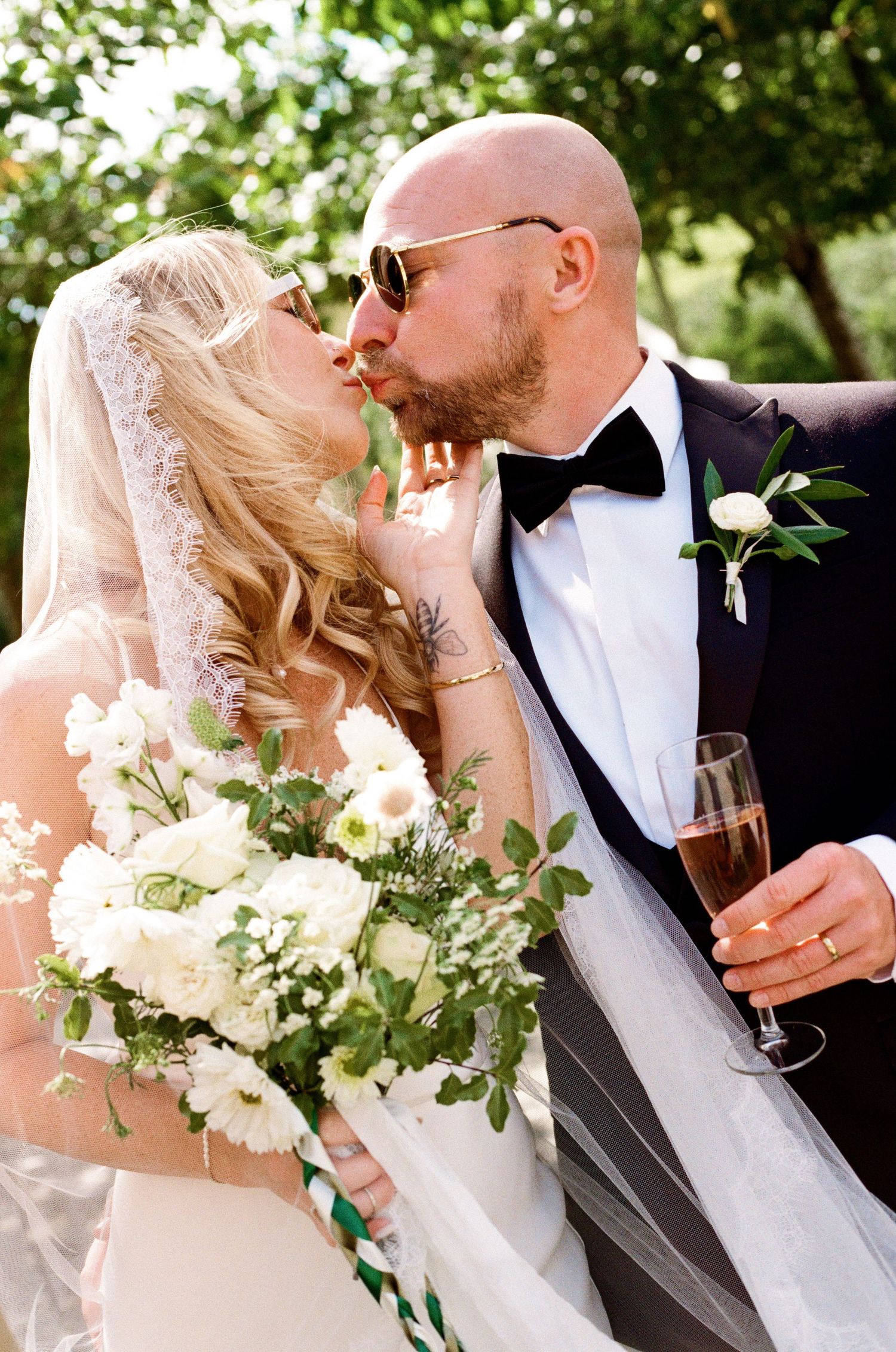 Wedding couple sharing champagne outdoors with elegant white floral bouquet.